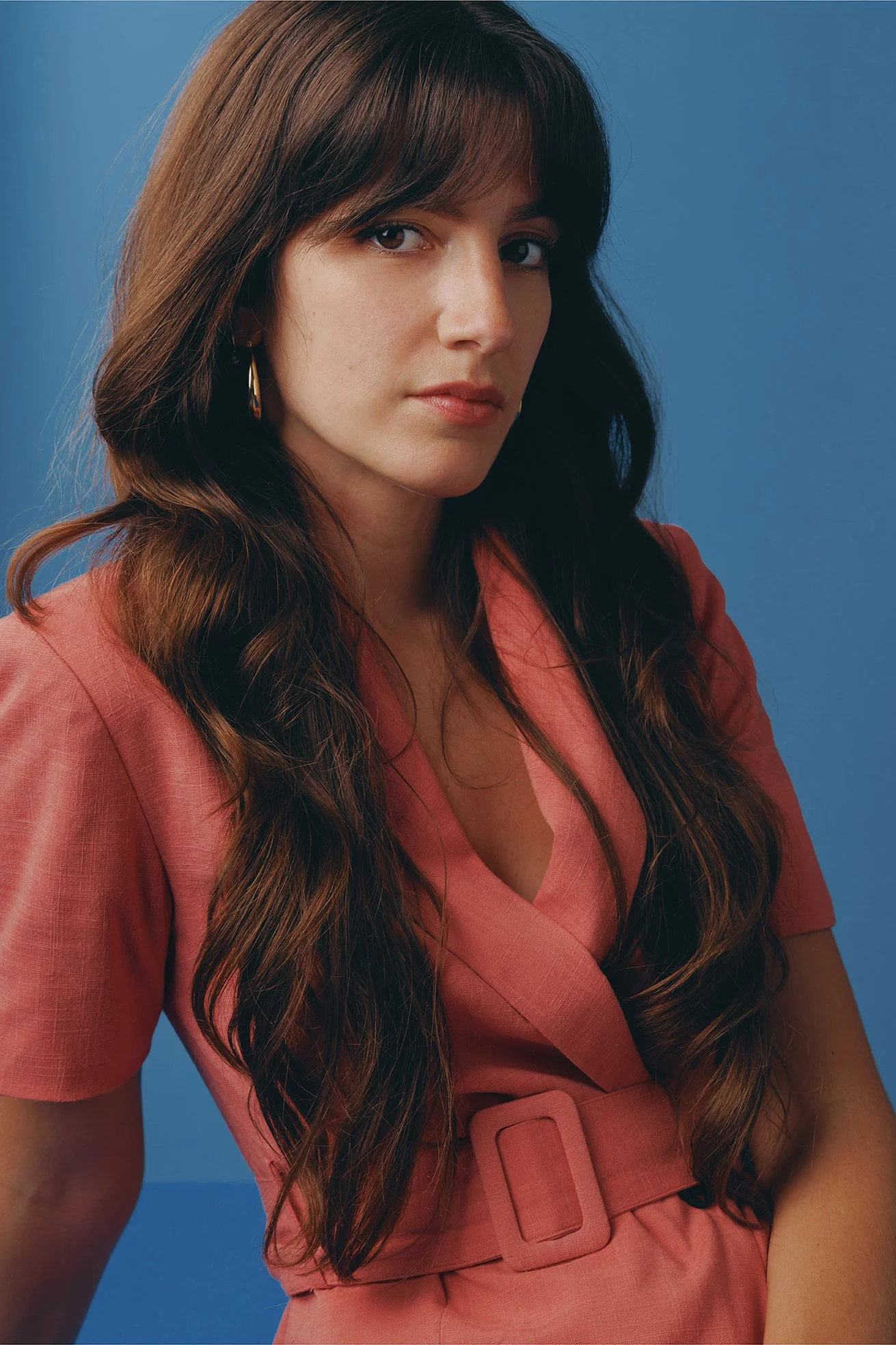 Close-up portrait of a woman with long, wavy dark hair, wearing a peach-colored blouse with a matching belt, standing against a solid blue background.