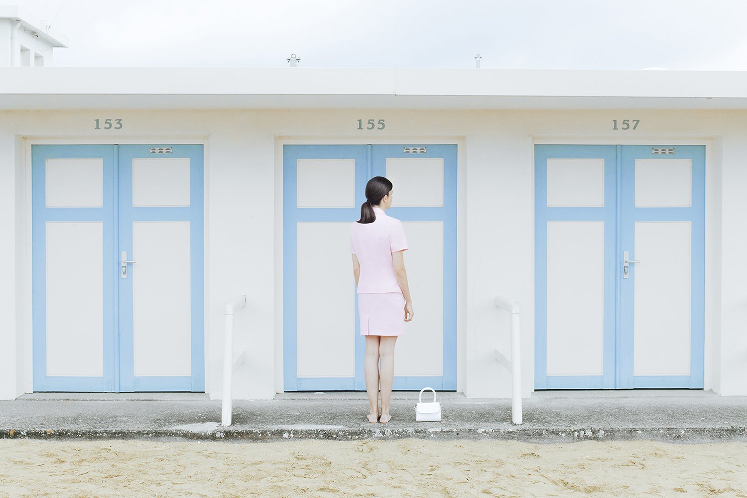 A woman in a pink dress standing in front of beach cabins with blue doors, numbered 153, 155, and 157, with a white handbag on the ground.