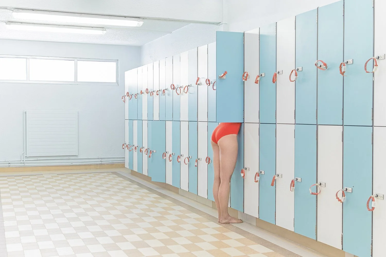 A person in orange swim shorts with their upper body inside a blue locker at a locker room, showing only their legs and lower torso.