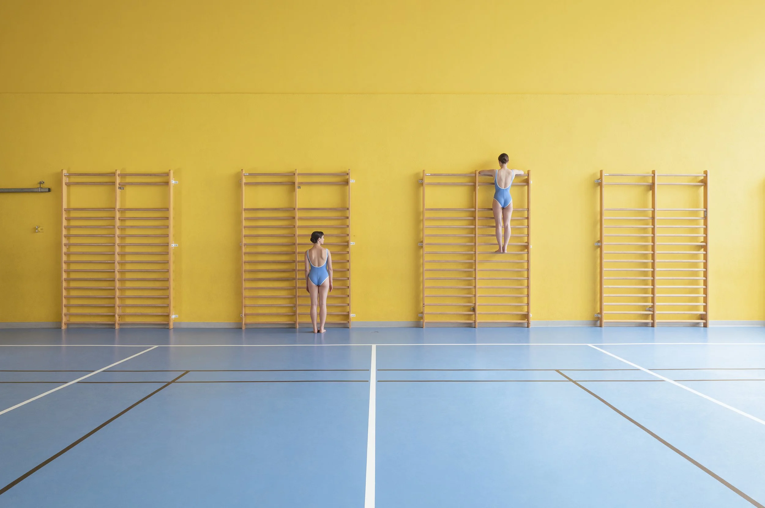 Two female gymnasts in blue leotards stretching on wooden wall bars in a gymnasium with a yellow wall, blue floor, and white lines.
