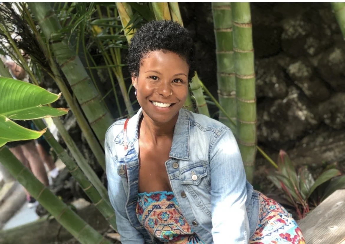 A woman with short curly hair and a bright smile, wearing a denim jacket and a colorful, patterned dress, sitting outdoors among tall bamboo plants.