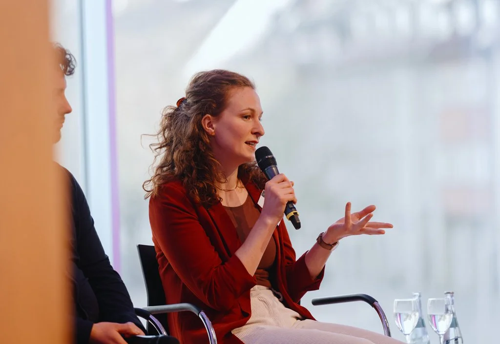 Leonie Pessara auf einer Bühne, spricht mit Mikrofon bei einer Podiumsdiskussion