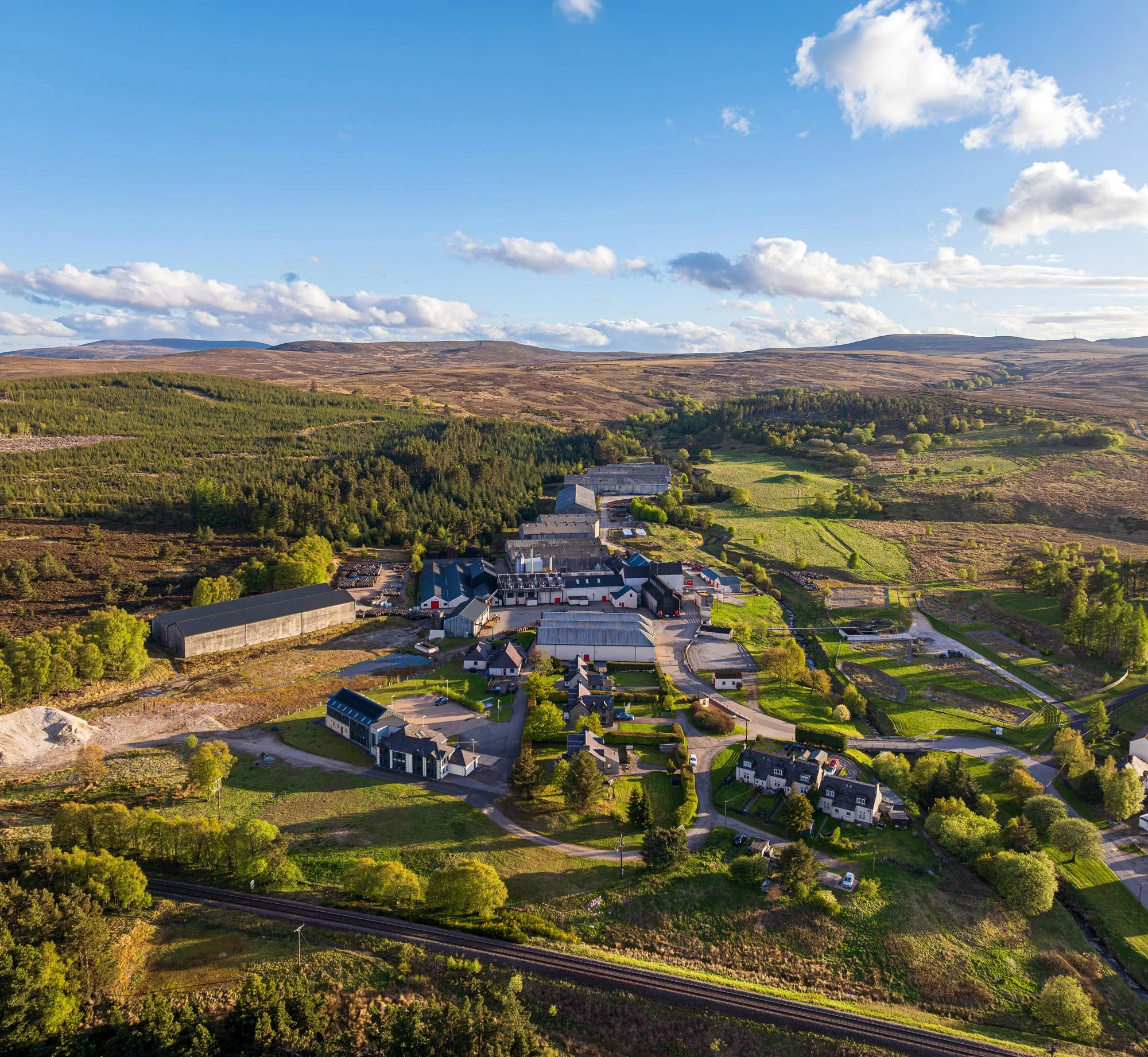 A rural landscape featuring a small village with homes and buildings, surrounded by green fields, forests, and rolling hills under a partly cloudy sky.