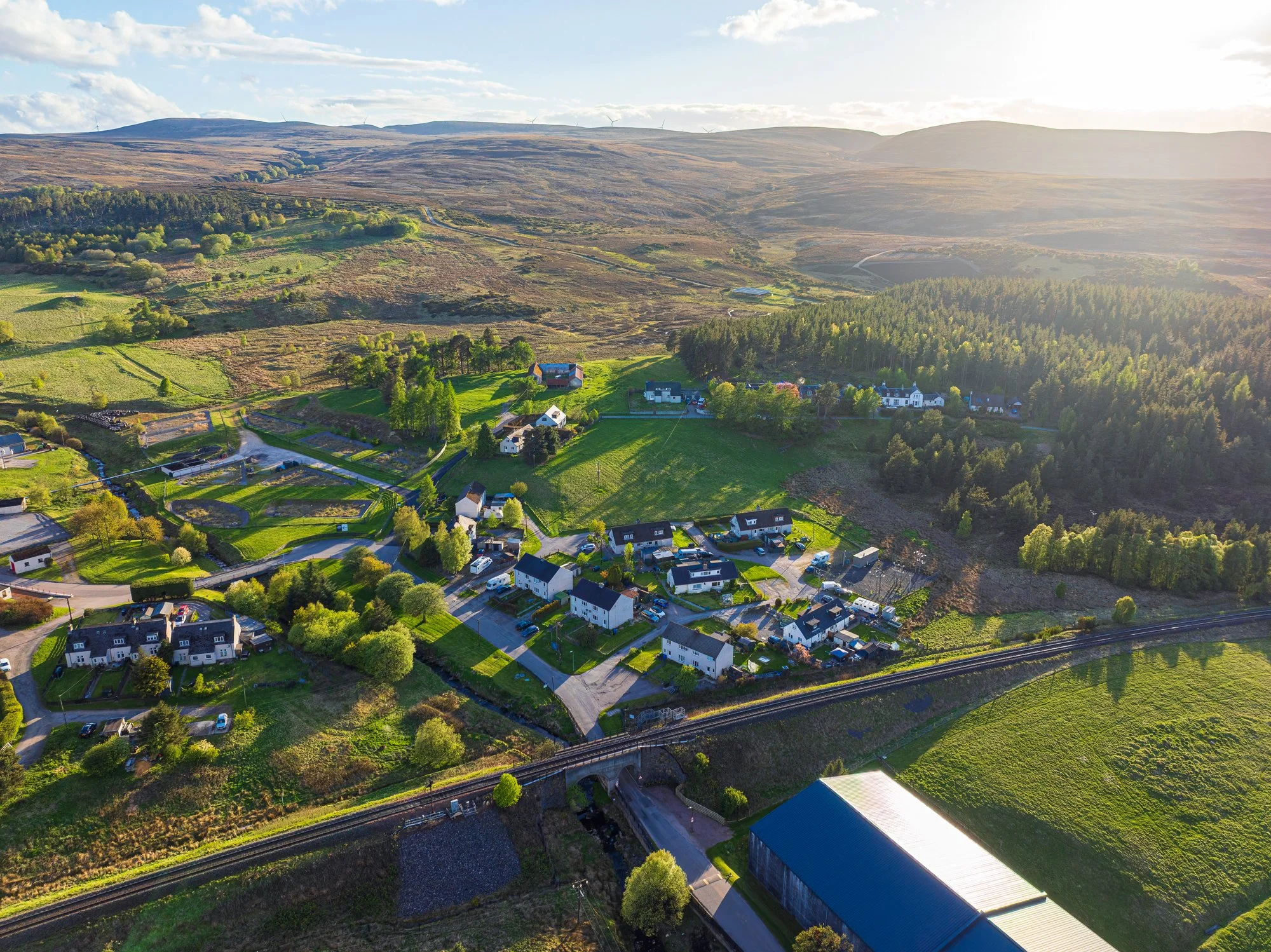 Aerial view of a rural landscape with houses, fields, trees, and a railway track with a bridge, surrounded by rolling hills and wind turbines in the distance.