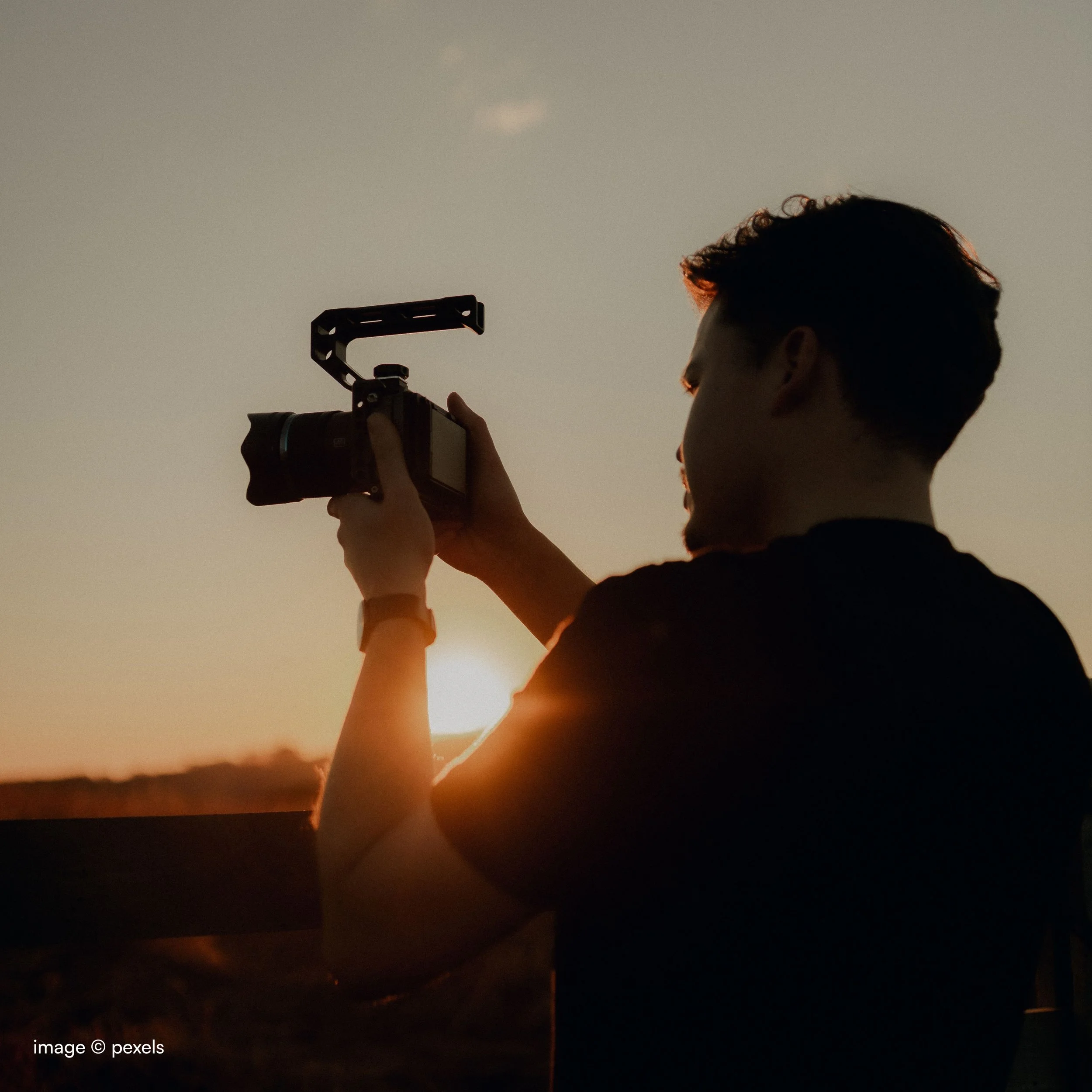 Person with short hair holding a camera during sunset, with the sun low on the horizon in a rural landscape.