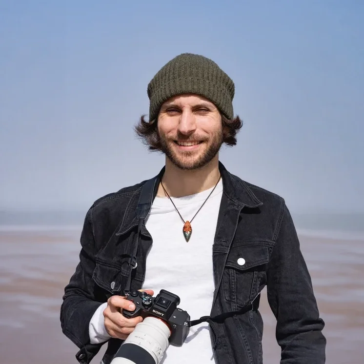 Smiling man outdoors on a beach, wearing a green knit beanie, black denim jacket, white shirt, and a necklace with a pendant, holding a camera.