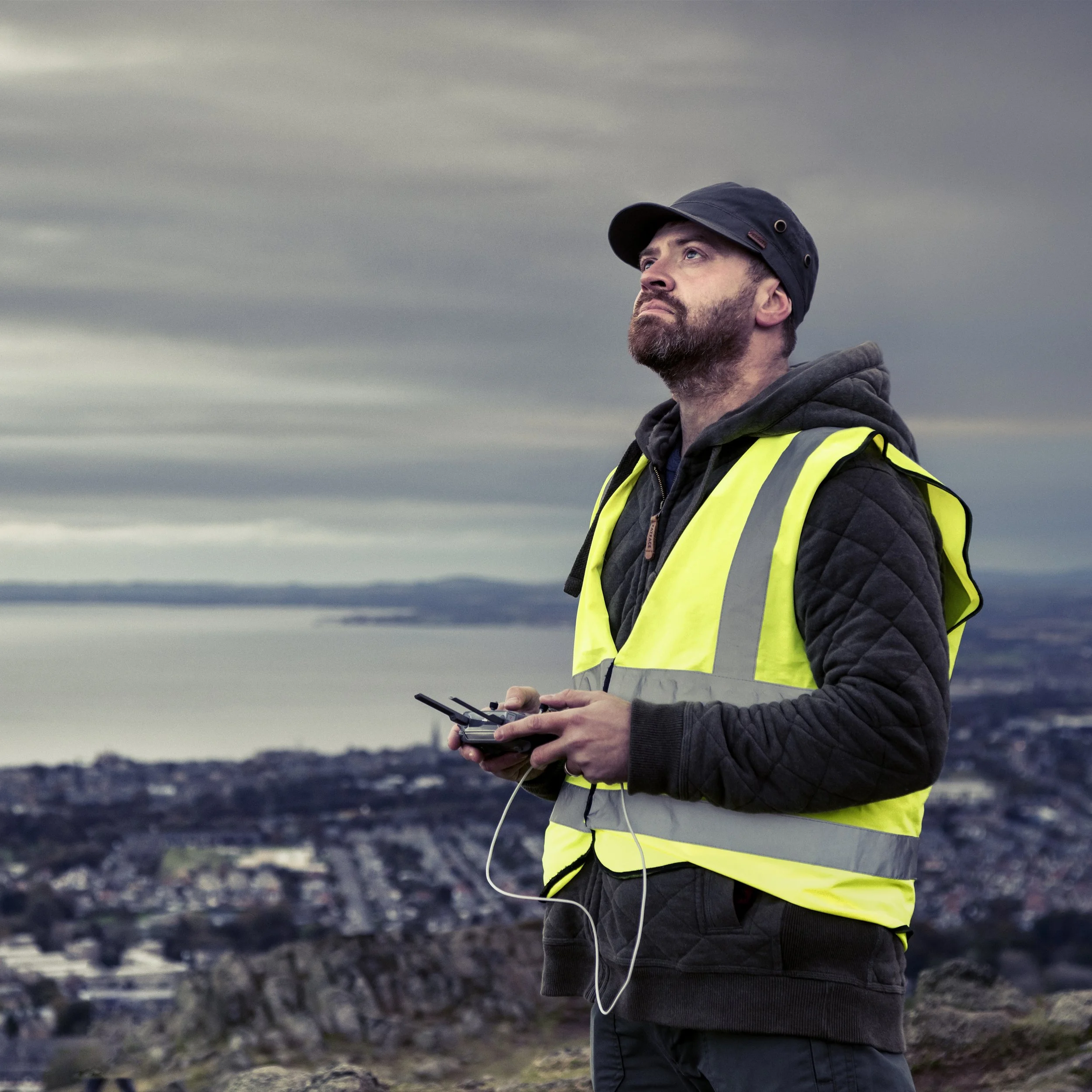 A man with a beard wearing a black cap, black jacket, and yellow safety vest is standing outdoors on a rocky trail. He is holding a remote control and looking up at the cloudy sky during dusk or dawn.