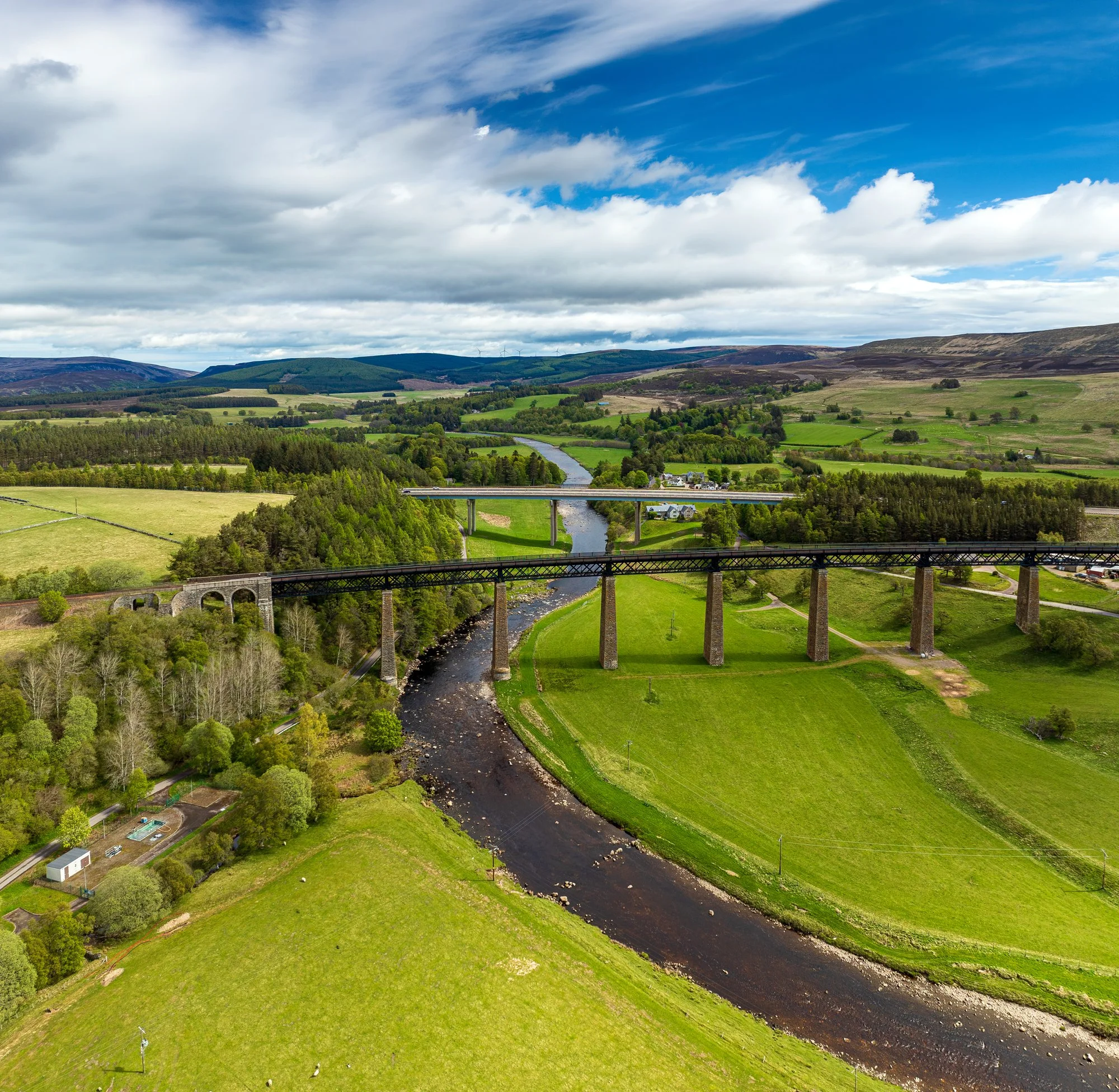 A landscape with rolling green hills, a river flowing through the valley, and a bridge crossing over the river. The sky is partly cloudy with some areas of blue sky visible.