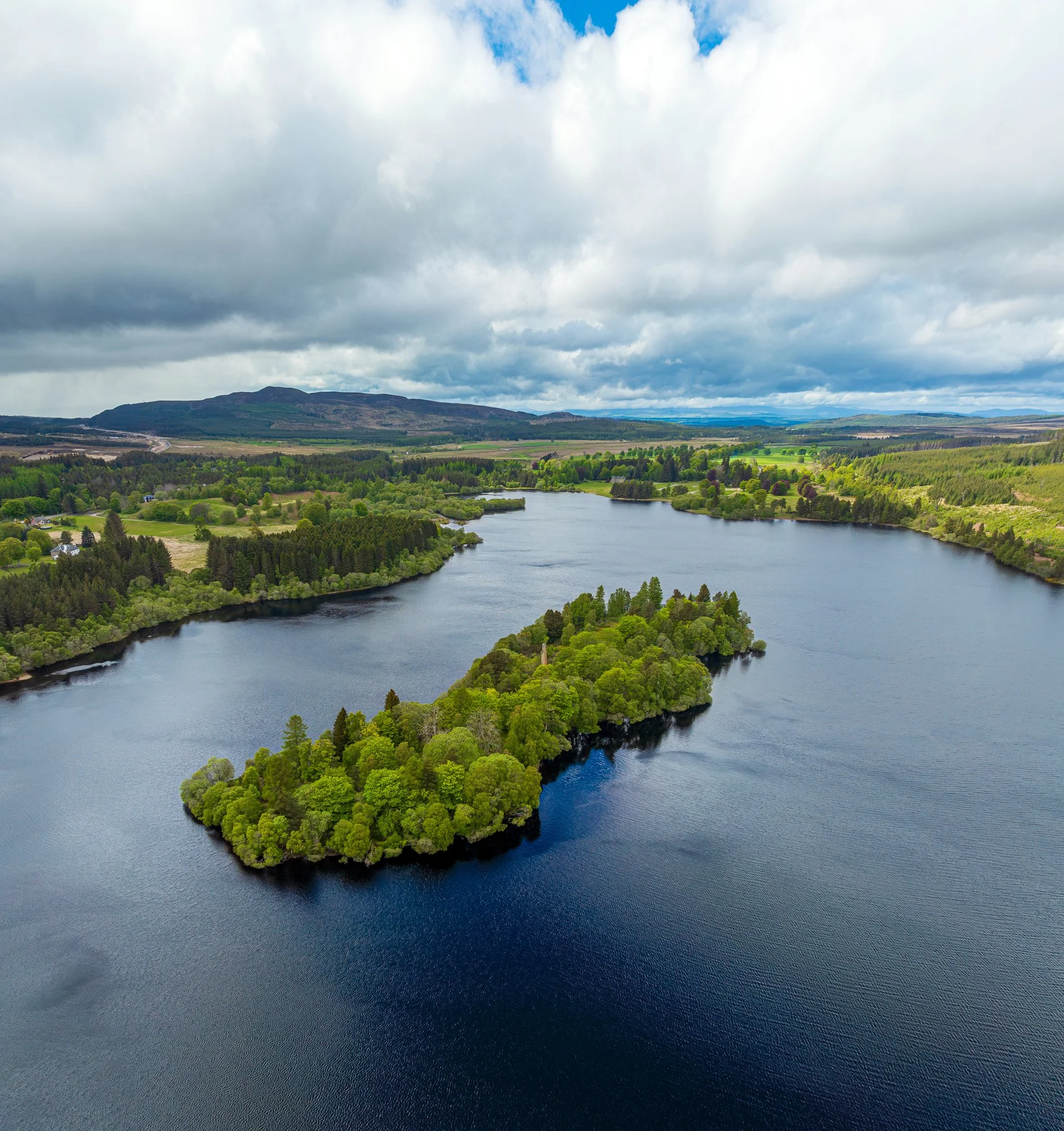 Aerial view of a river meandering through lush green trees and countryside with hills and cloudy sky in the background.