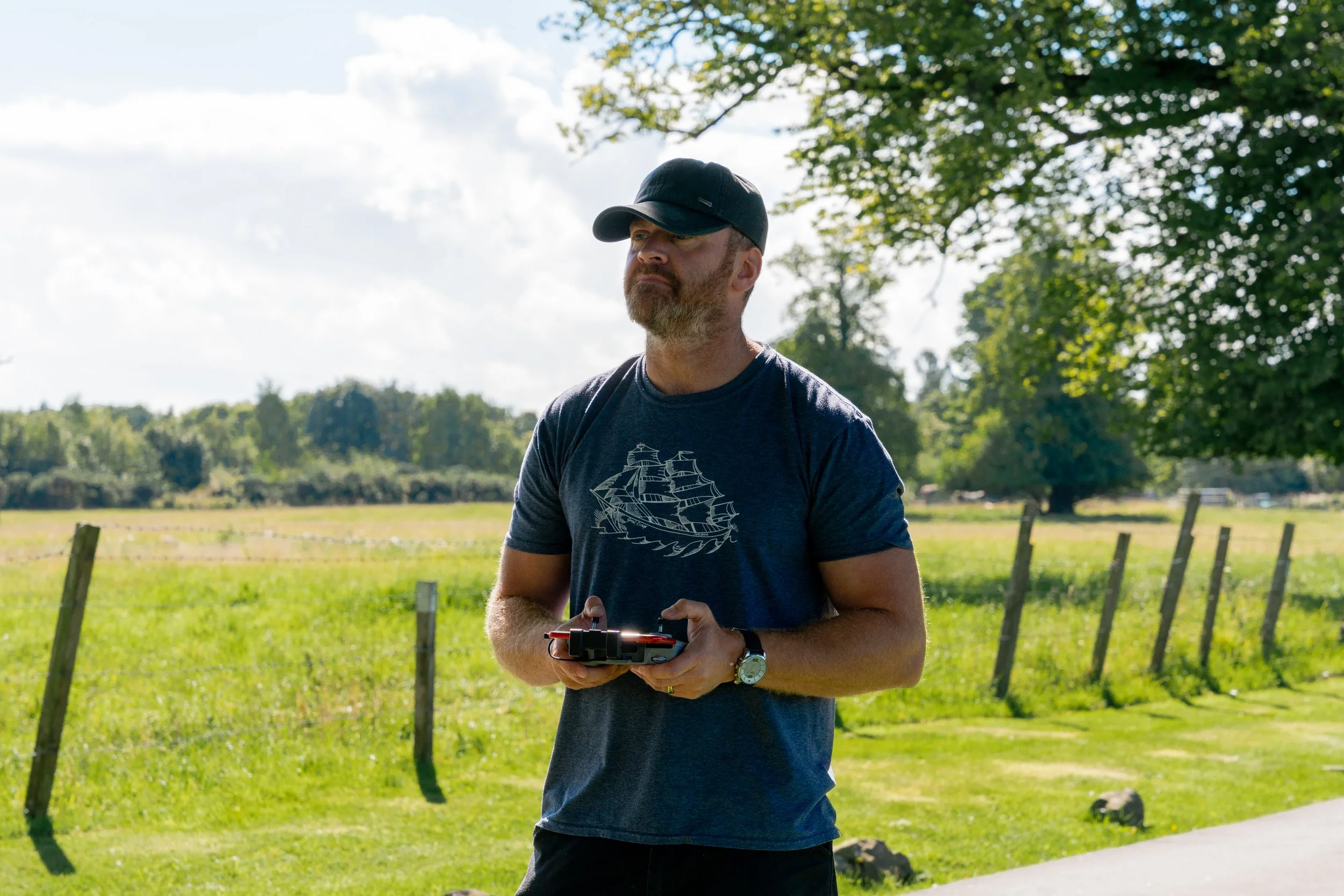 A man with a beard and mustache wearing a black cap, navy t-shirt with a ship print, and a watch on his left wrist, holding a remote control, standing outdoors in a green field with trees and a blue sky with clouds in the background.