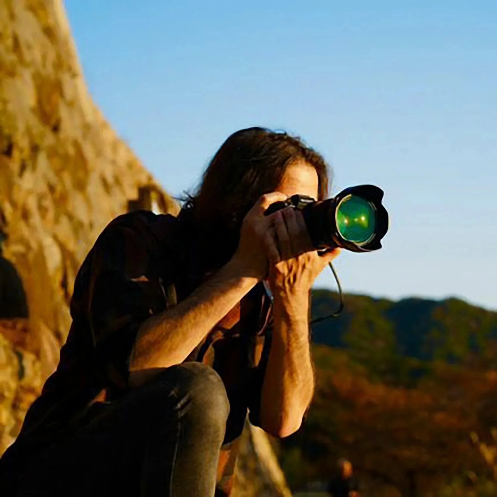 A man with dark hair, wearing a black shirt, sitting outdoors at night, looking through a camera or binoculars, with a stone wall to her left and a dark evening sky in the background.
