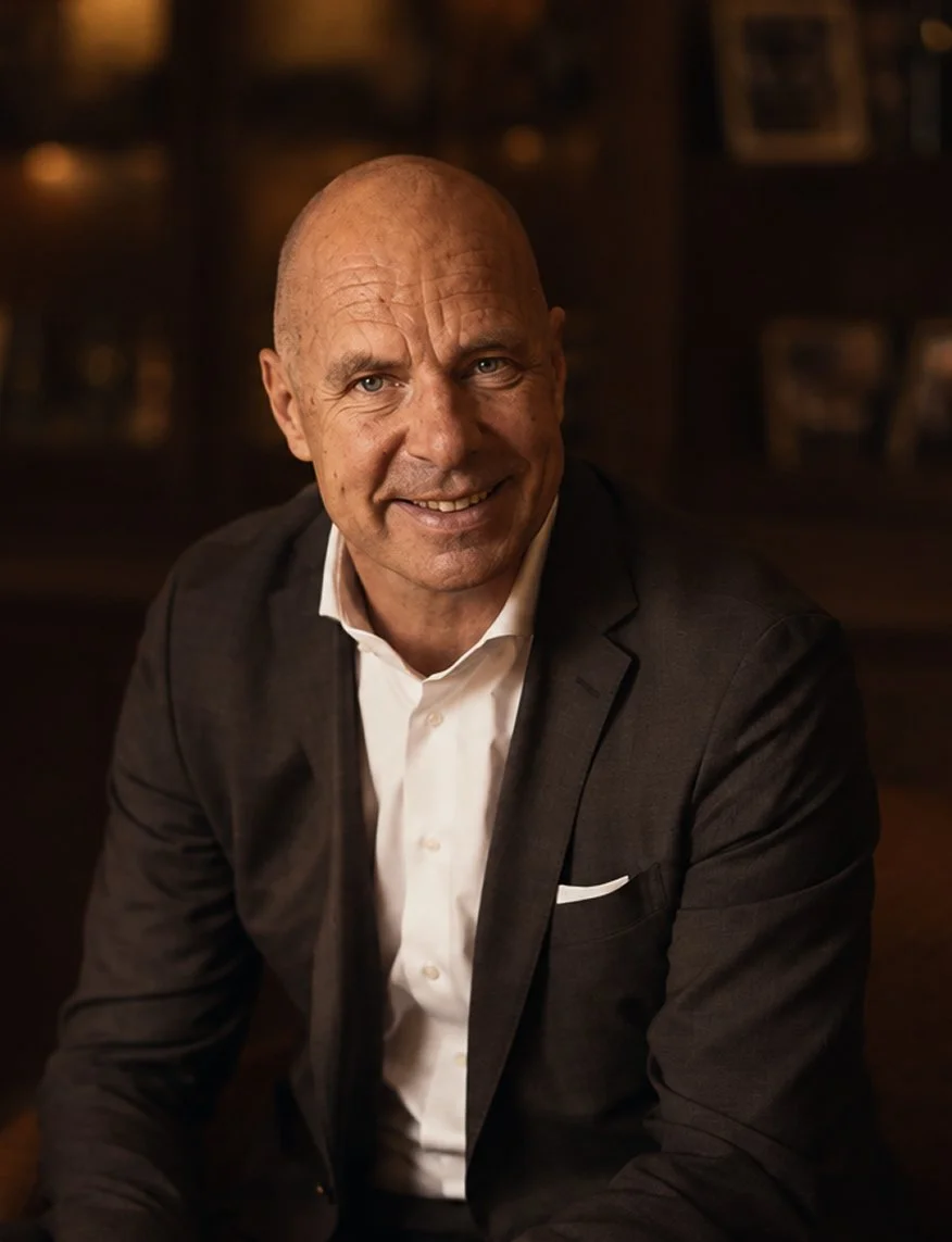 A middle-aged man with a bald head, smiling, wearing a dark suit jacket and white shirt, sitting in a dimly lit room with a blurred background.
