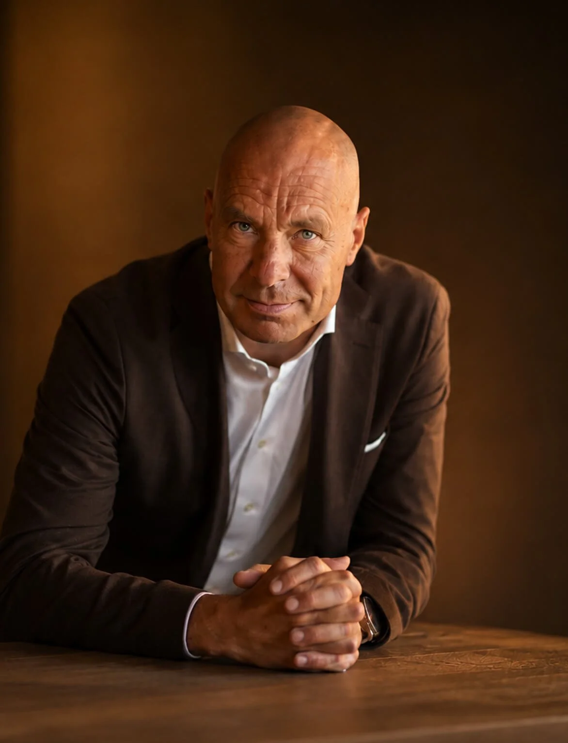 A bald man in a brown blazer and white shirt sitting at a wooden table with hands clasped, looking at the camera with a slight smile. The background is a solid dark color.
