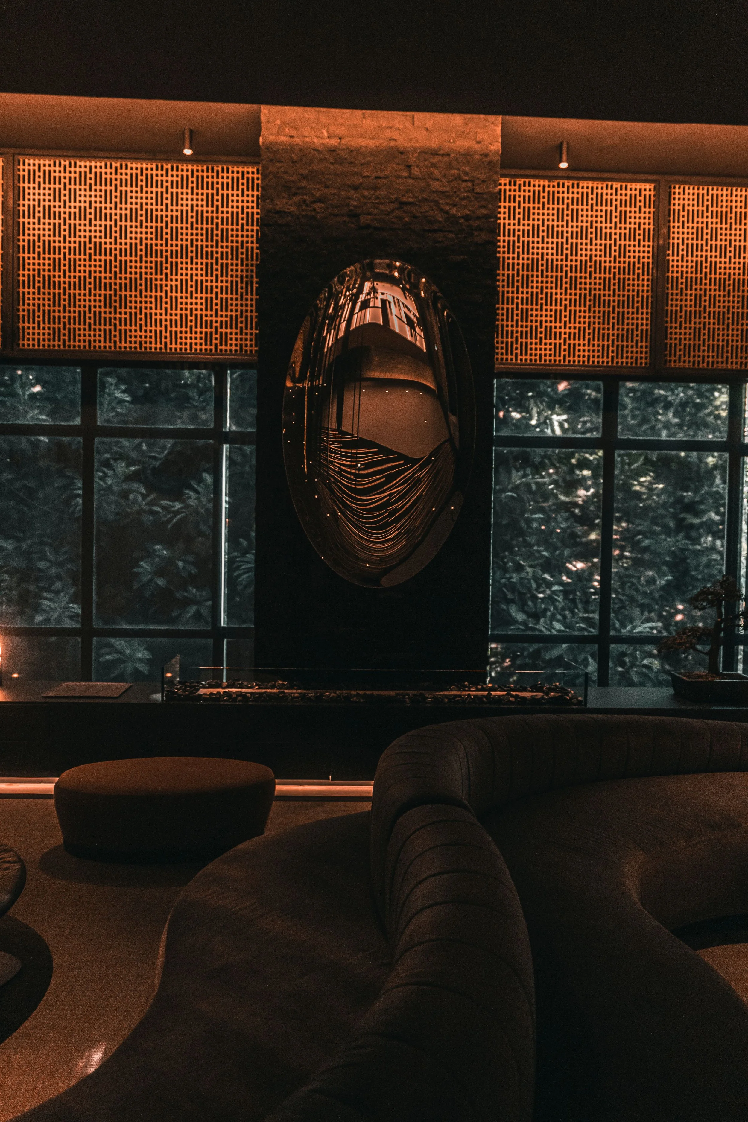 Interior of a modern lounge with dark walls, large windows, a curved gray sofa, a small table, and a unique oval-shaped decorative mirror reflecting the room, illuminated with warm ambient lighting.