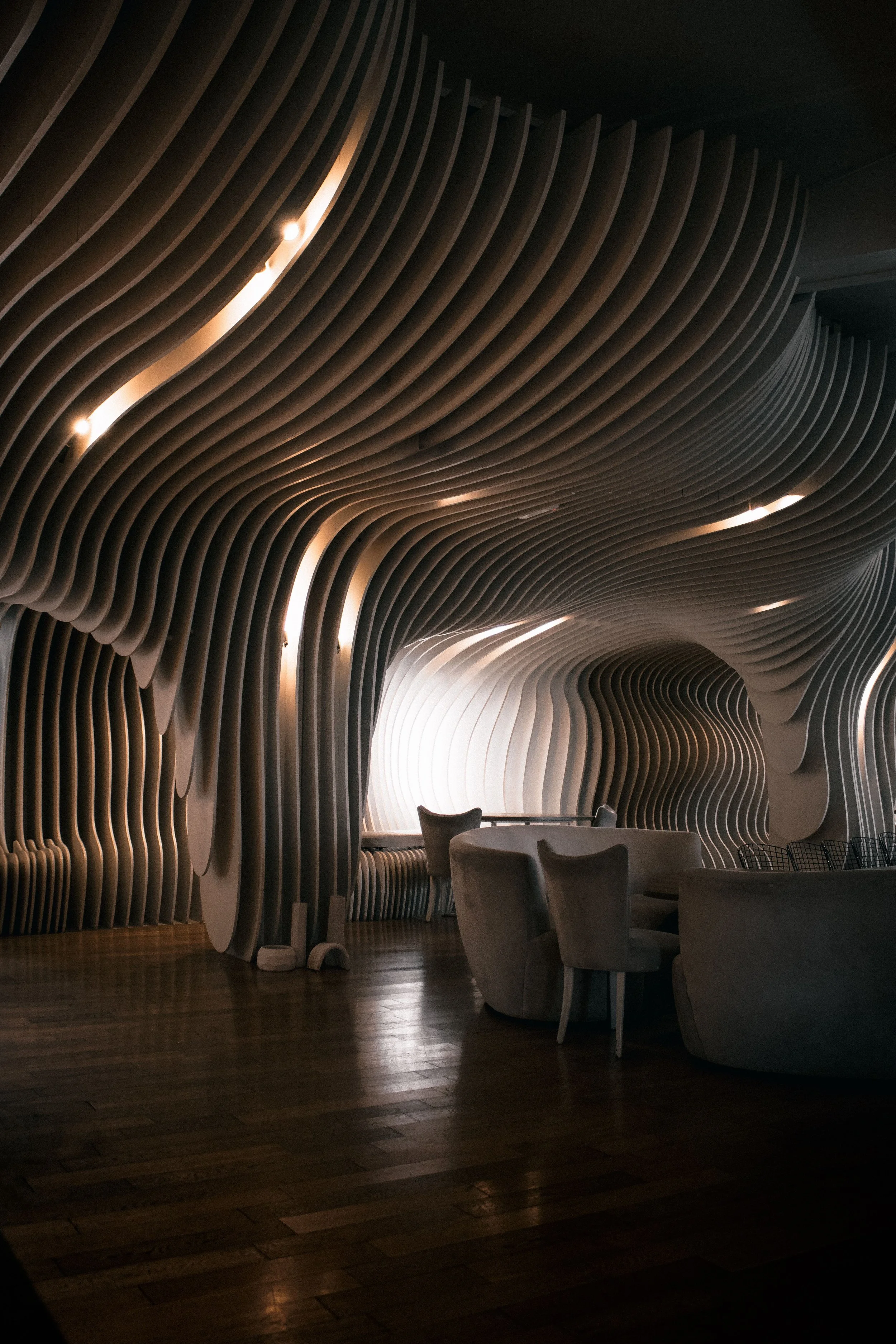 A modern interior space featuring a ceiling with curved, layered wooden slats and built-in lighting. There are chairs and a table in the foreground, with a dark wooden floor.
