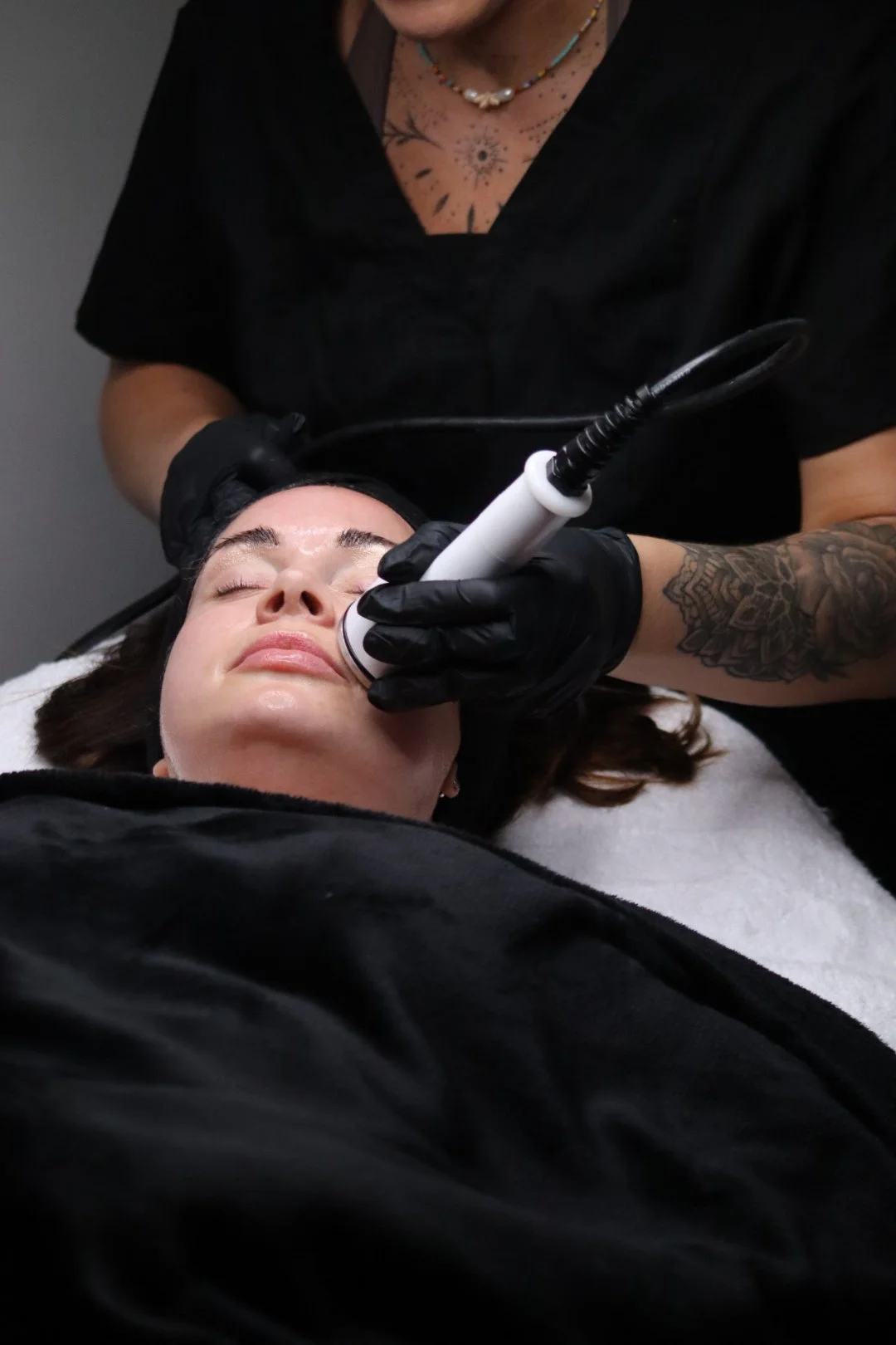 A woman receiving a facial treatment with a handheld device from a professional wearing black gloves in a spa or clinic setting.