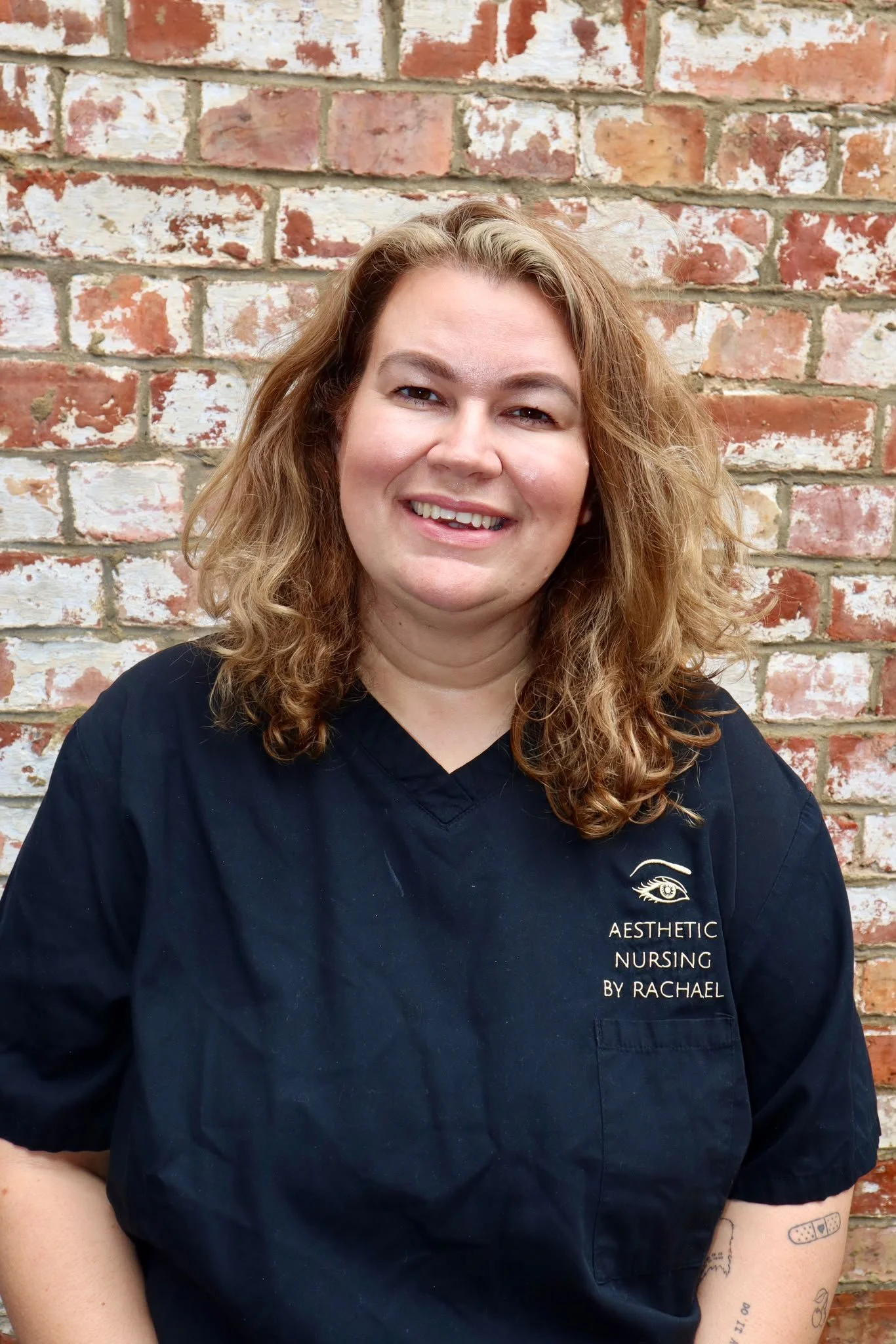 A woman with curly hair smiling in a black medical uniform, standing in front of a framed art print of a syringe and lipstick.