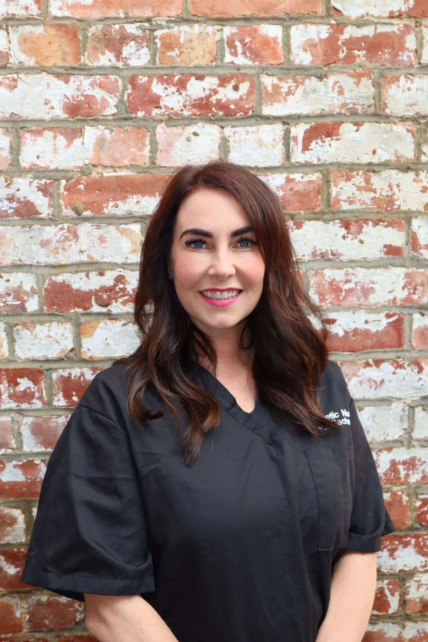 A woman with long brown hair, blue eyes, and pink lipstick is smiling and wearing a black shirt, standing in front of a red and white brick wall.