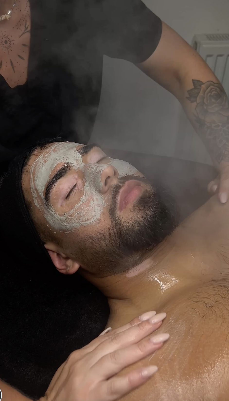 Man with a beard lying down with a facial mask applied, receiving a relaxing facial treatment in a spa setting.