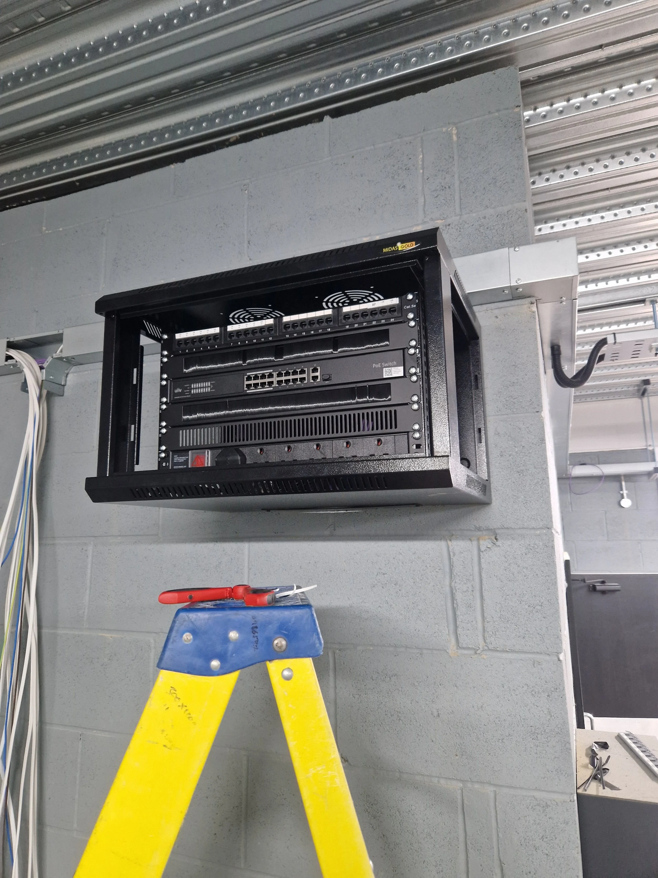 Electrical rack mounted on a gray wall with yellow ladder in front of it, orange-handled tool on top of the ladder, various wires and tools nearby.