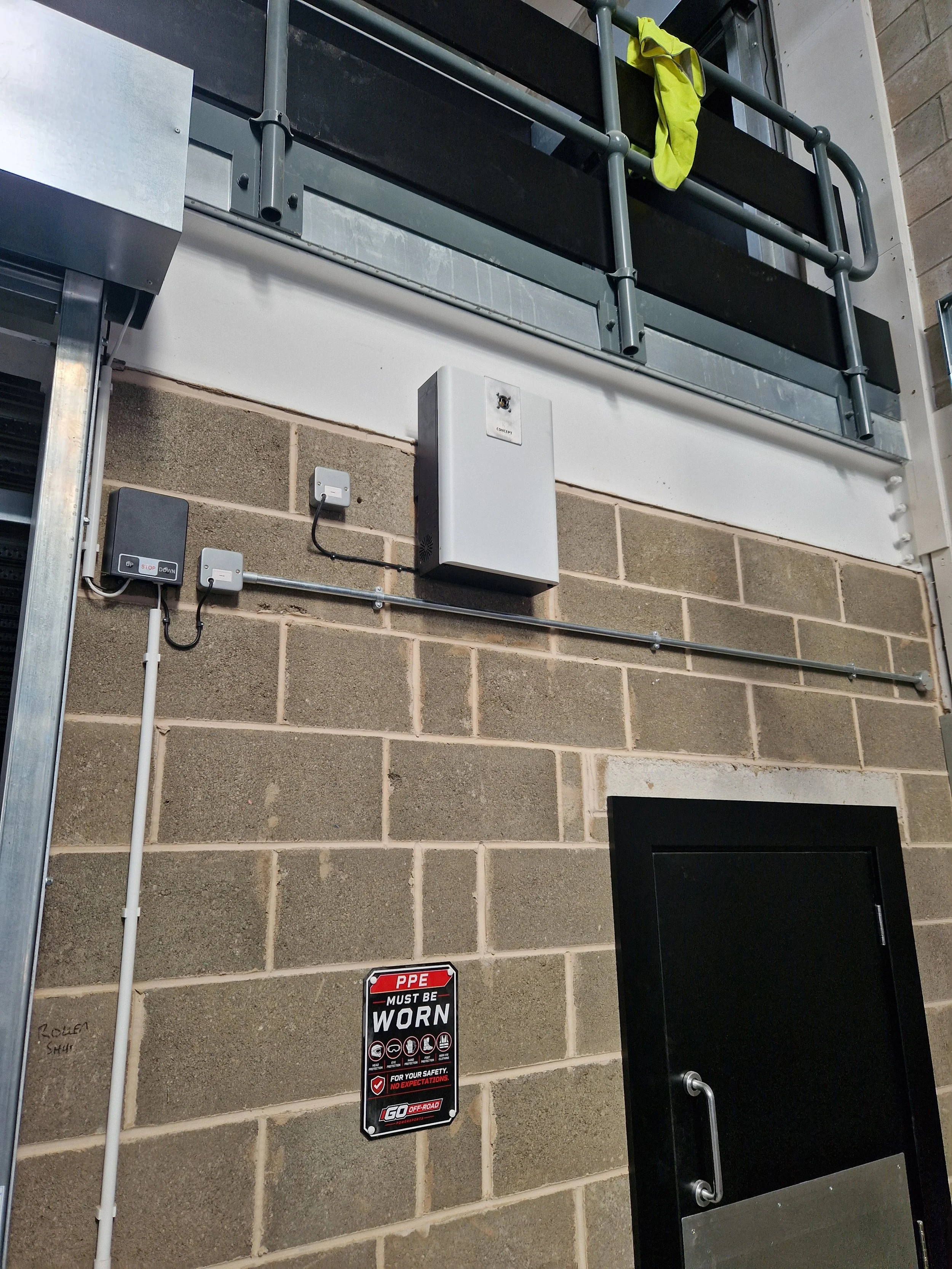 Close-up of a building wall with electrical equipment, including a box labeled 'CONCEPT', wiring, an electrical panel, and safety signs, including PPE must be worn.