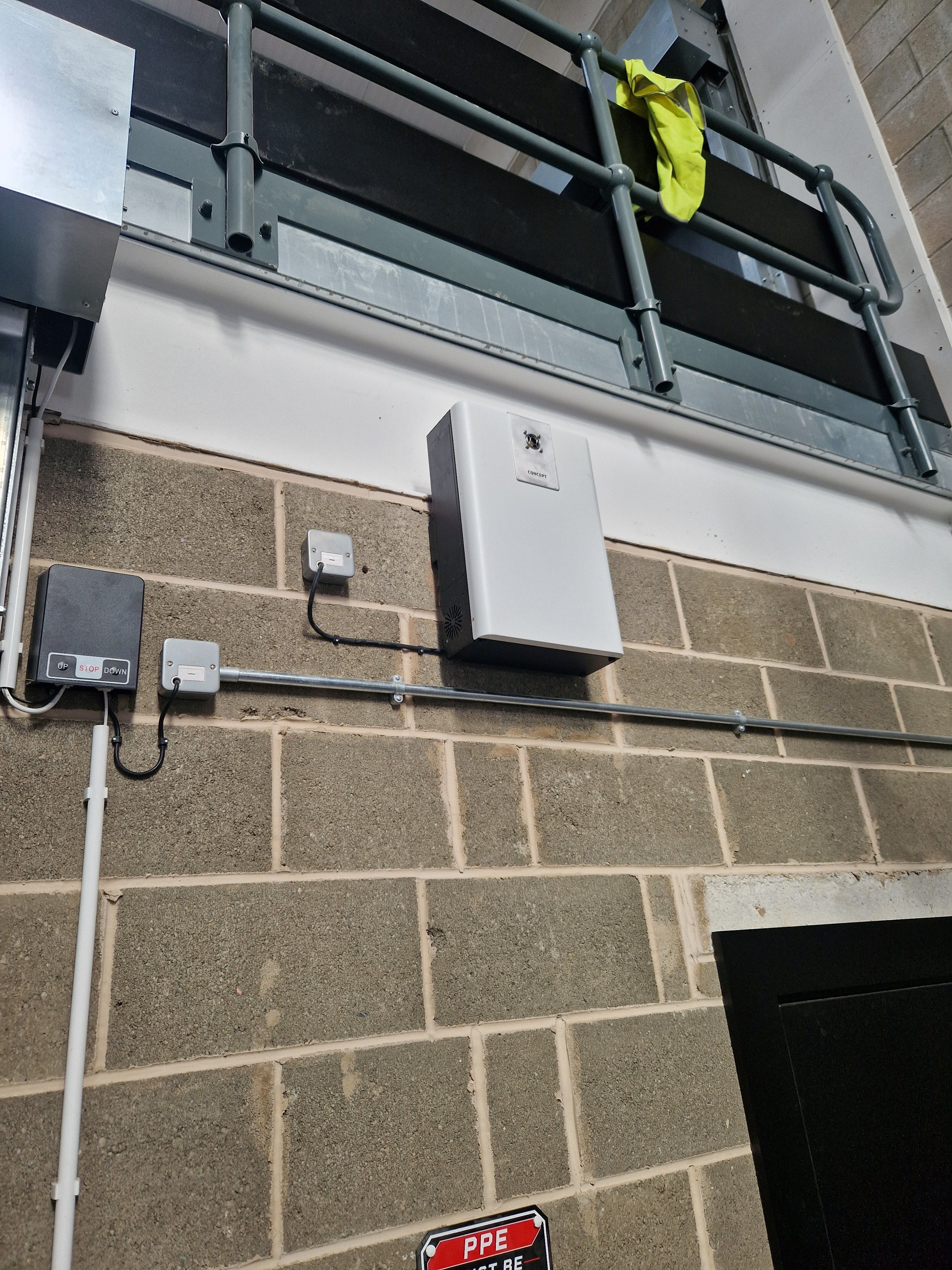 Close-up of security equipment on a brick wall, including a black and gray electronic access control keypad, a white box with a black speaker grille, and a rectangular sign with red, black, and white colors that says "PPE" and "MUST BE WORN".