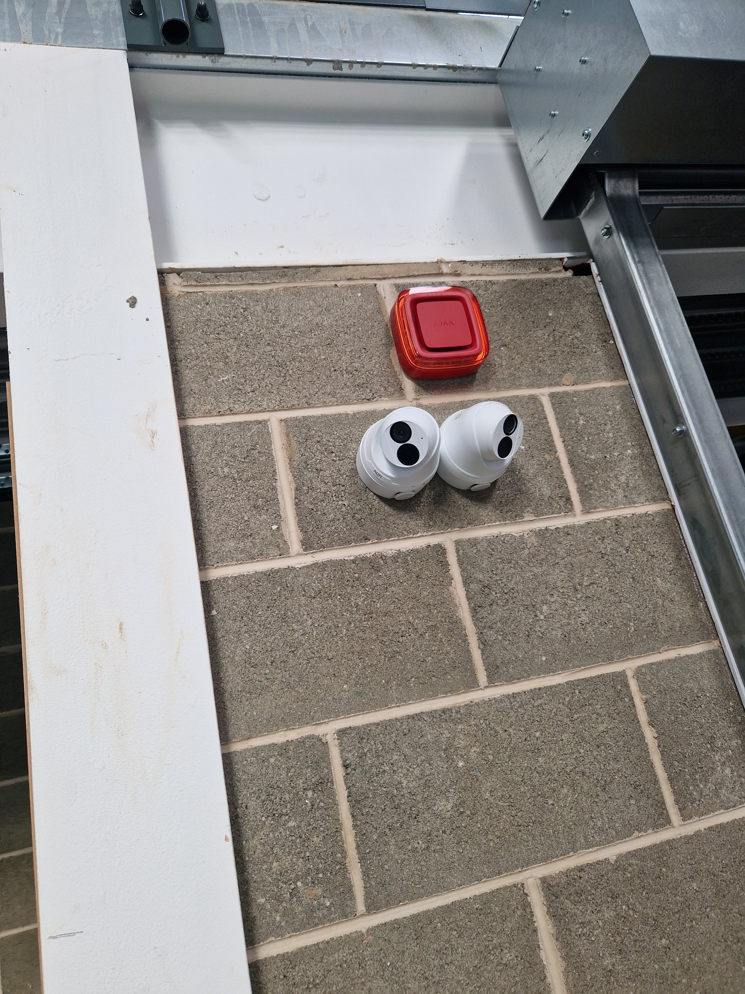 Two security cameras and a red alarm box are mounted on a brick wall near the ceiling, with parts of a metal frame and electrical wiring visible.