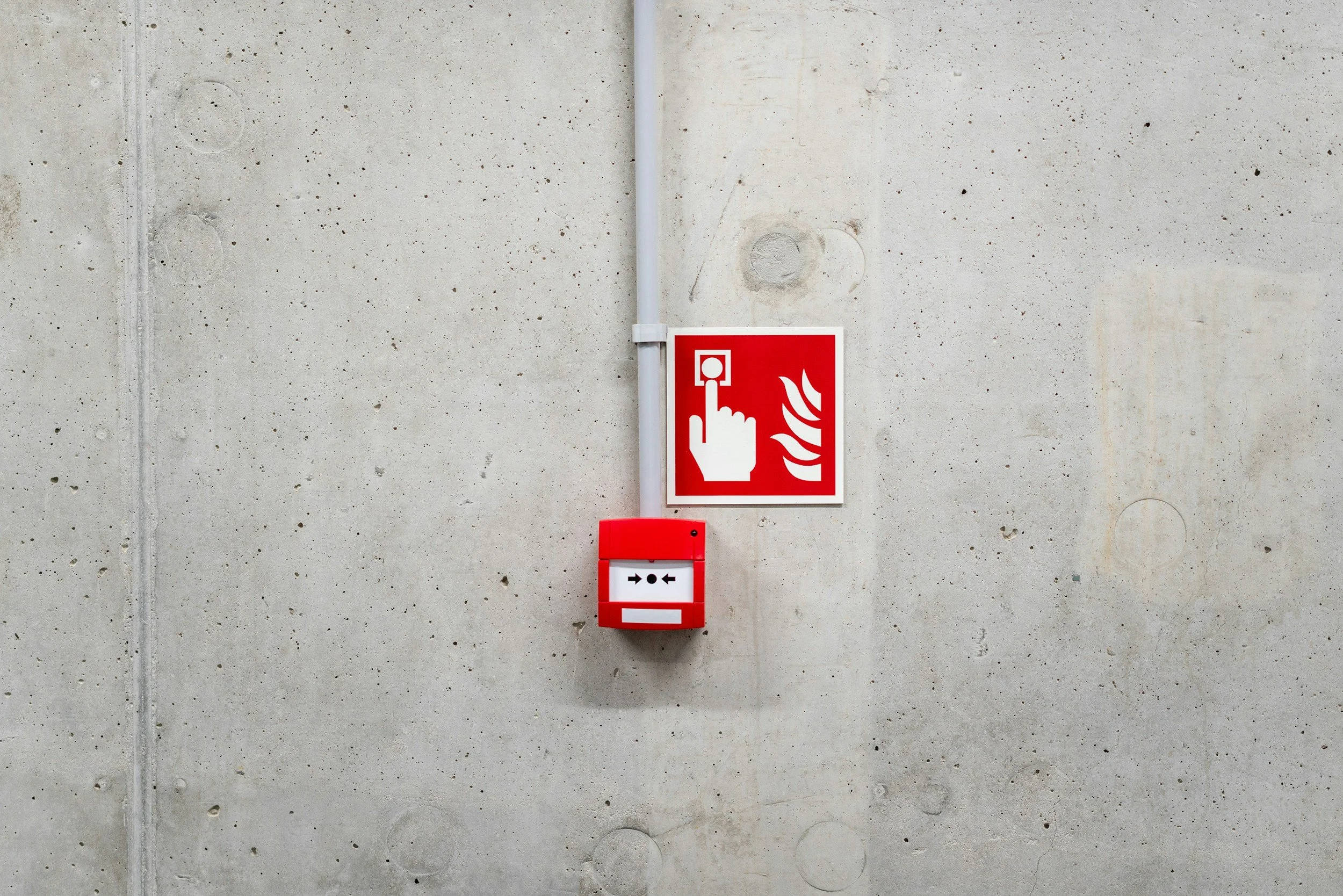 A red fire alarm pull station on a concrete wall with a white vertical pipe and a red sign with a hand pressing a button.
