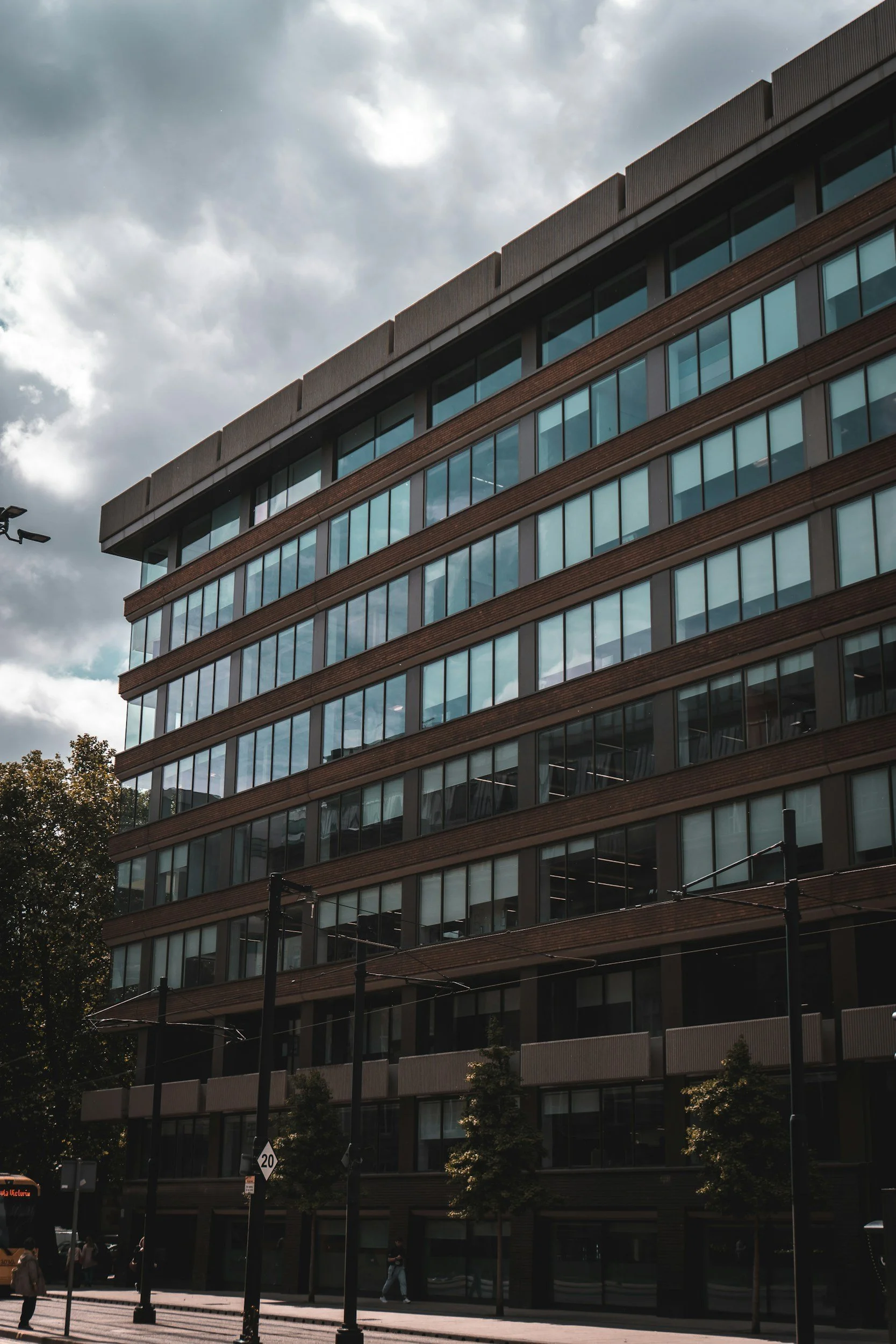 A tall, modern building with multiple floors and large glass windows, set against a cloudy sky. There are trees and street lamps in the foreground, with people walking on the sidewalk.