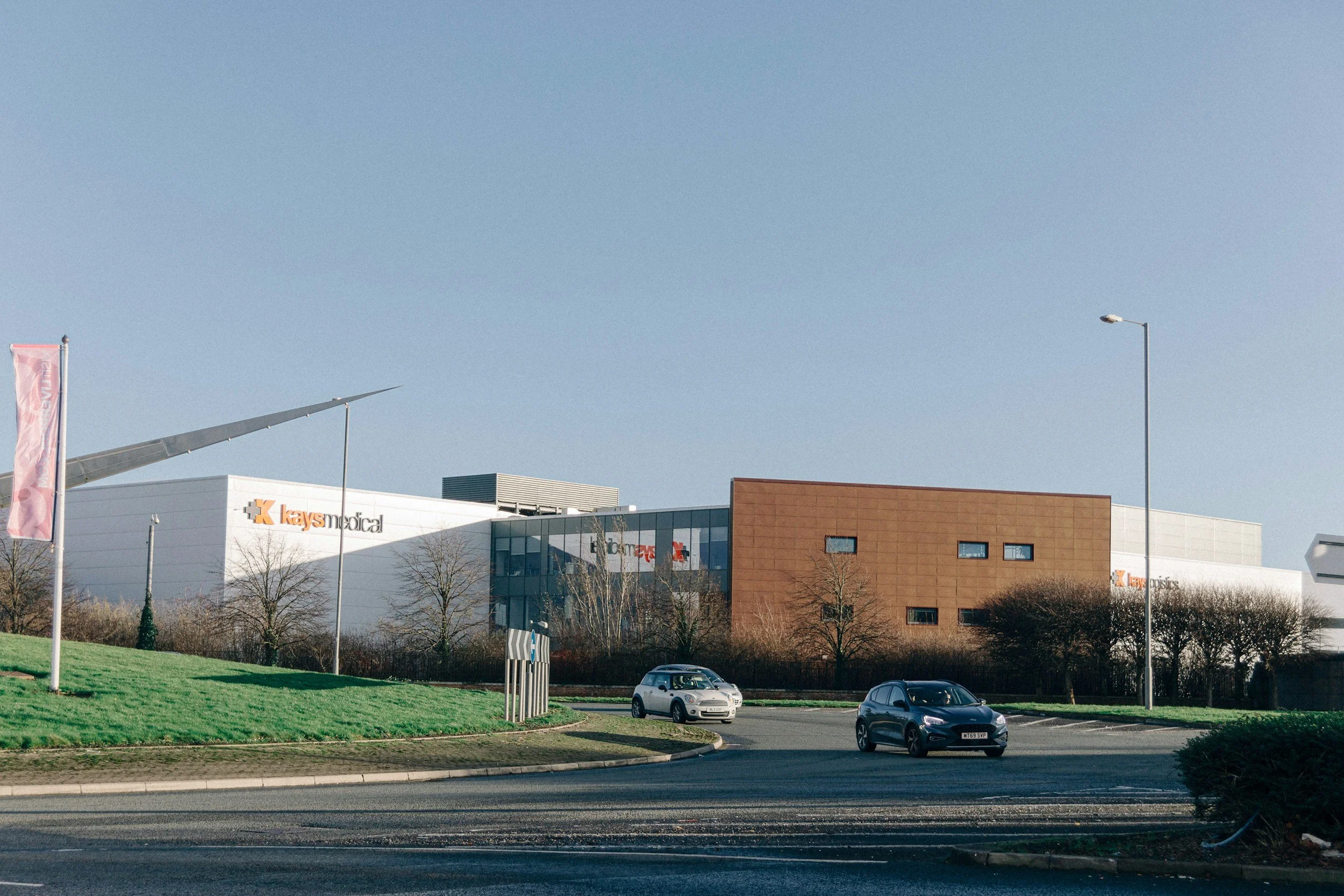 Parking lot with a few cars in front of a commercial building, under a clear blue sky. The building has signs for 'Kays Medical' and 'Kays Poses'.