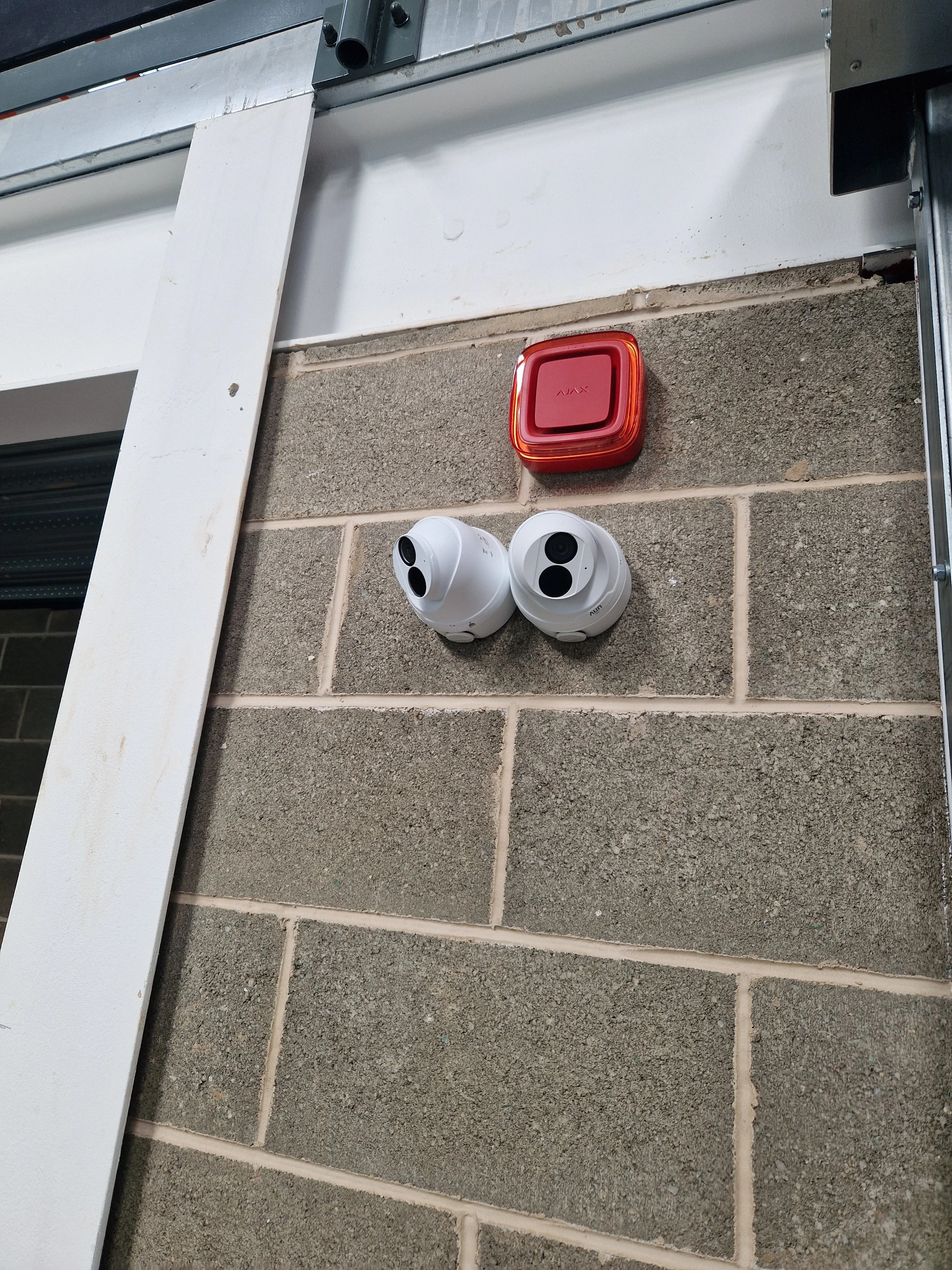 A red alarm button and two white security cameras mounted on a brick wall near a metal frame and a white painted board.