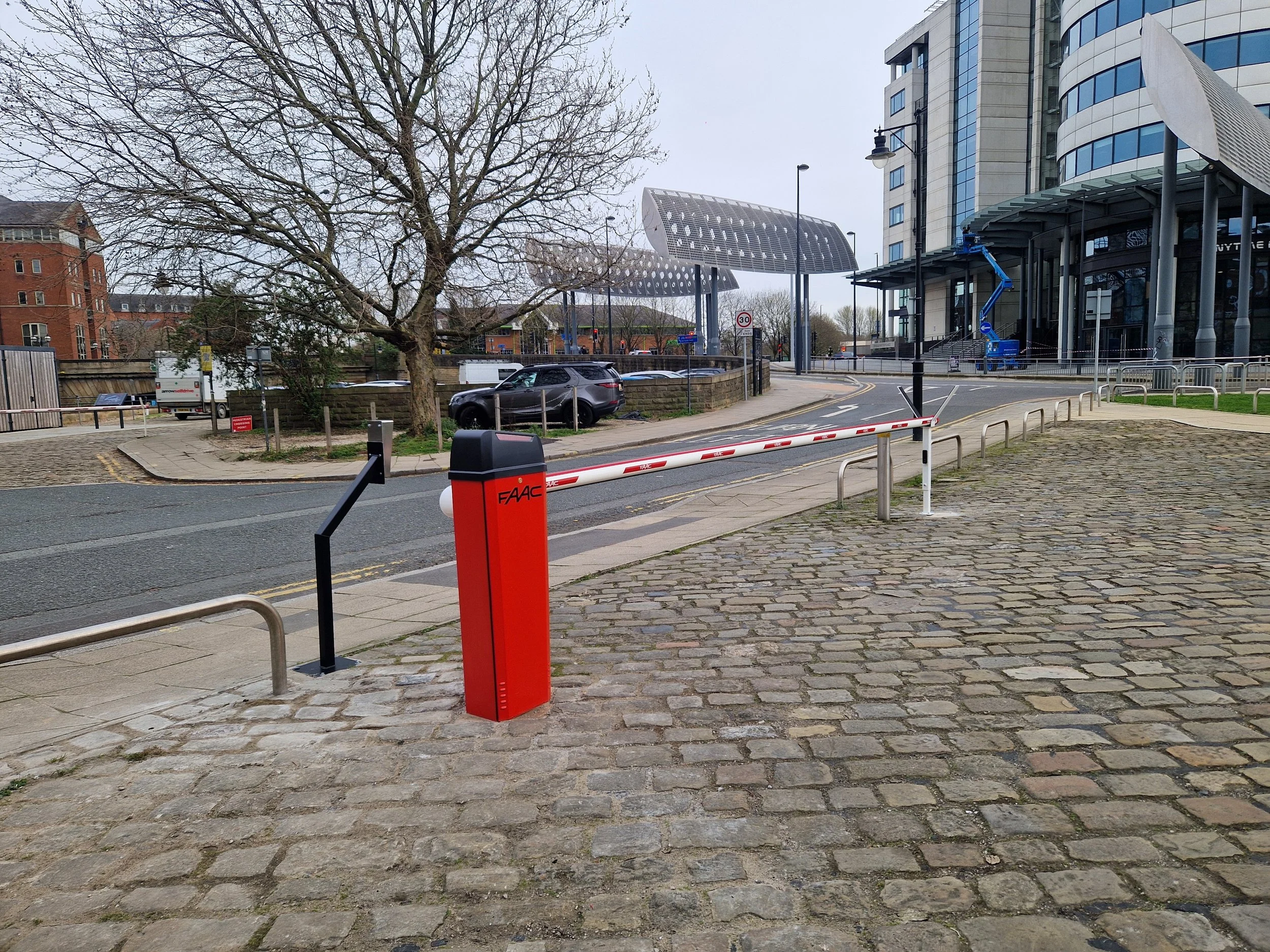 City street scene with leafless trees, modern glass office buildings, parked cars, and a red and white barrier gate.