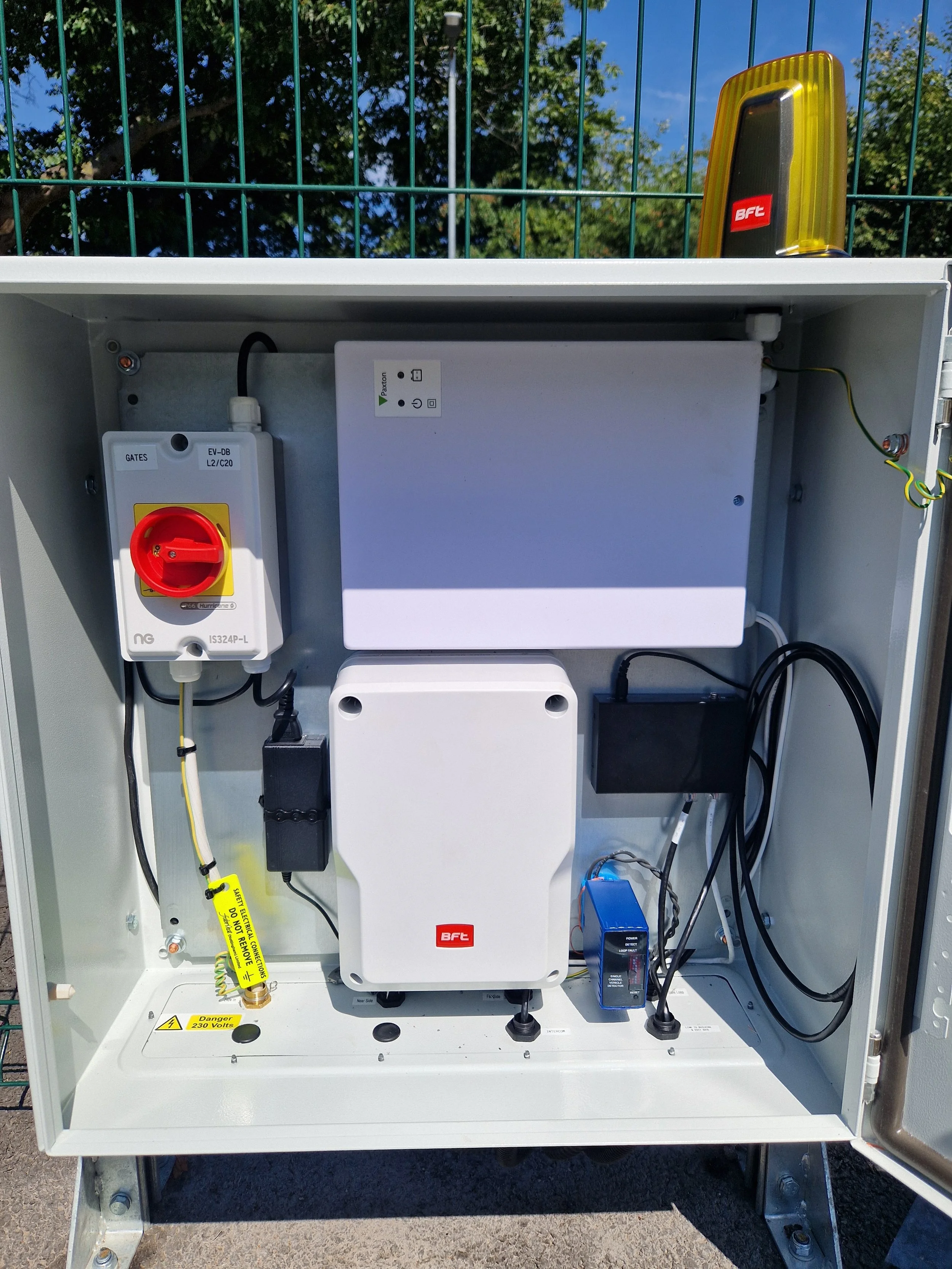 Open electrical cabinet containing various electrical components, wires, and a yellow warning sticker, with trees and a blue sky in the background.