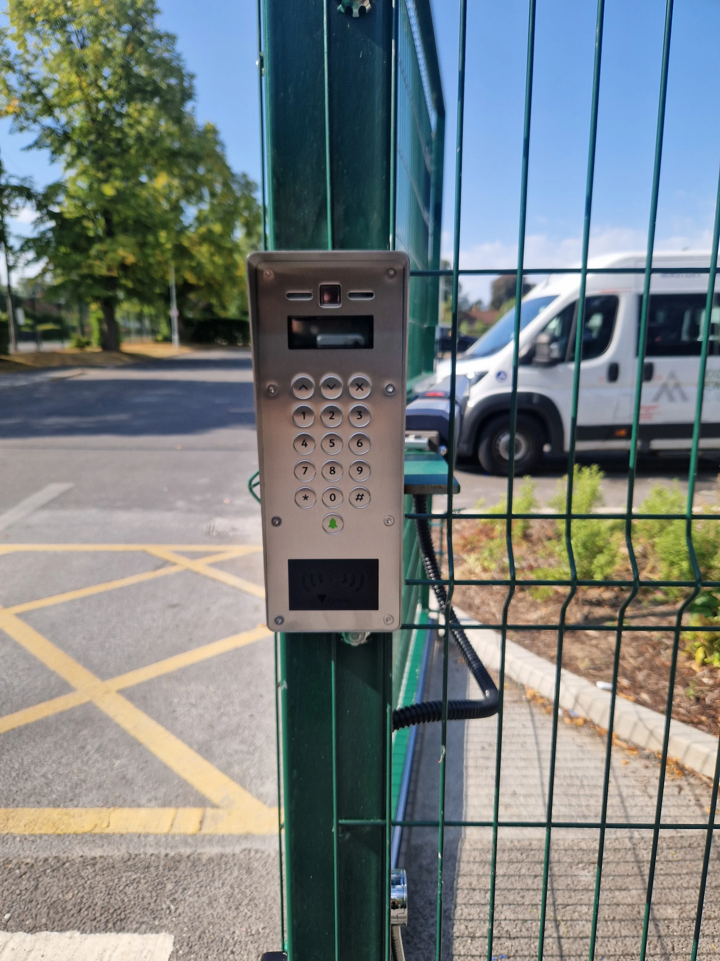 Access control keypad and card reader attached to a green metal fence, with a parking lot and trees visible in the background.