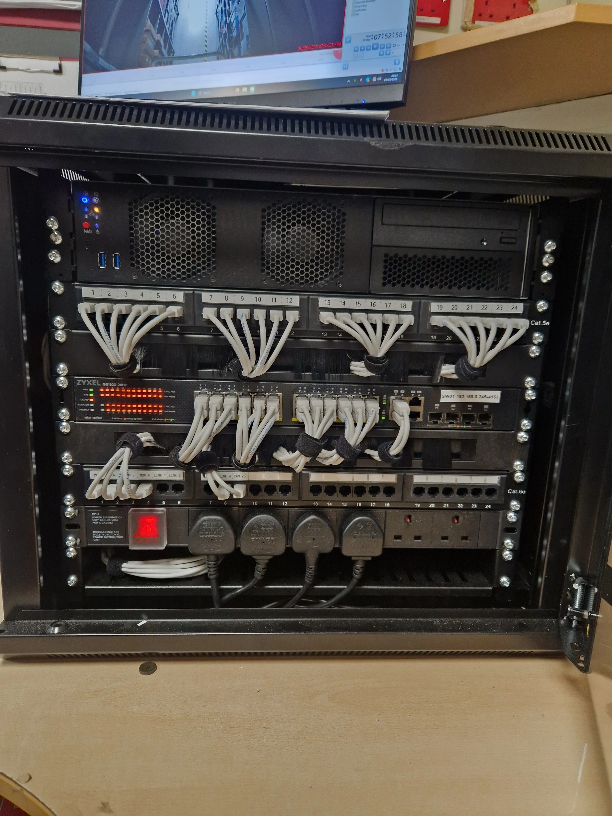 A server rack mounted in a metal case with network cables connected to various ports, a power distribution unit, and a monitor displaying surveillance footage on top of the rack.