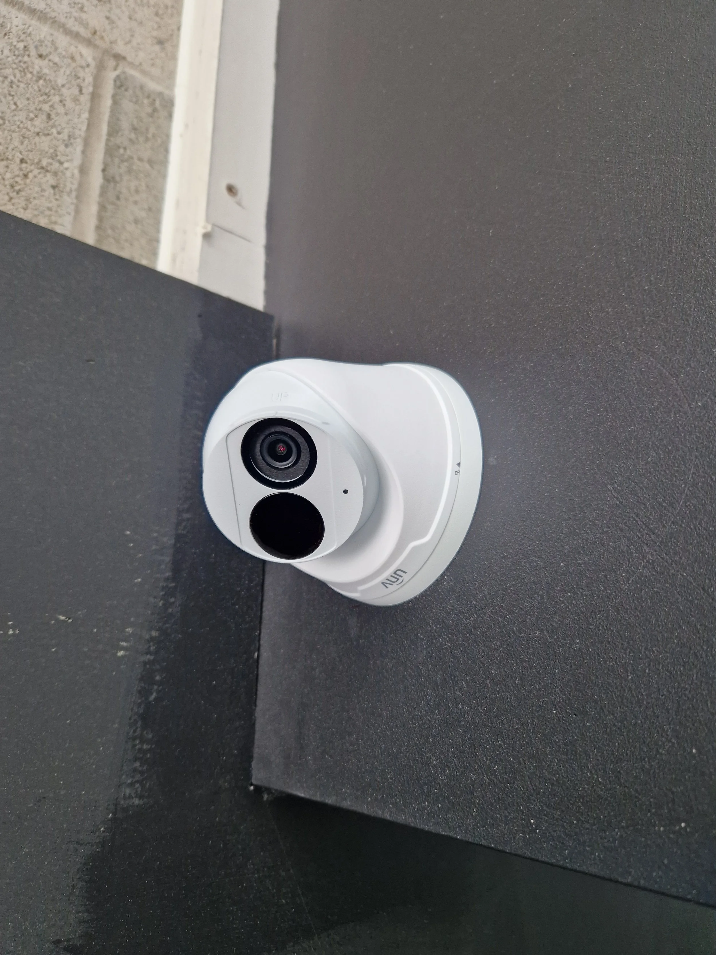 A white security camera mounted on a black surface beneath a siding wall.