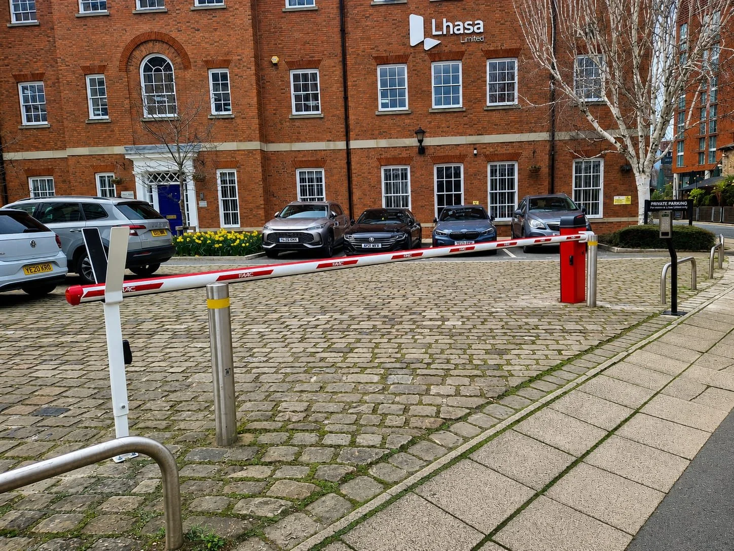Parked cars behind a barrier in front of a brick building with a sign that reads 'Lhasa Limited.' There are trees and a sidewalk on the right side.