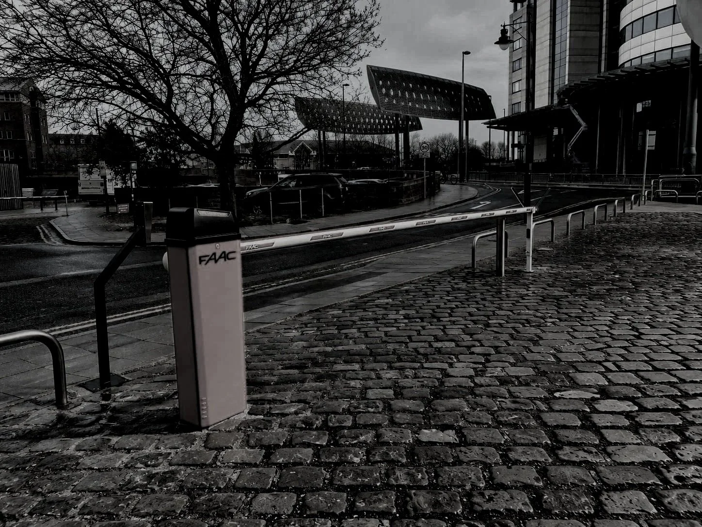 Empty urban street scene in black and white, showing a Cobblestone sidewalk, a parking barrier with FAAC written on it, parked cars, a leafless tree, modern buildings, street lamps, and a cloudy sky.