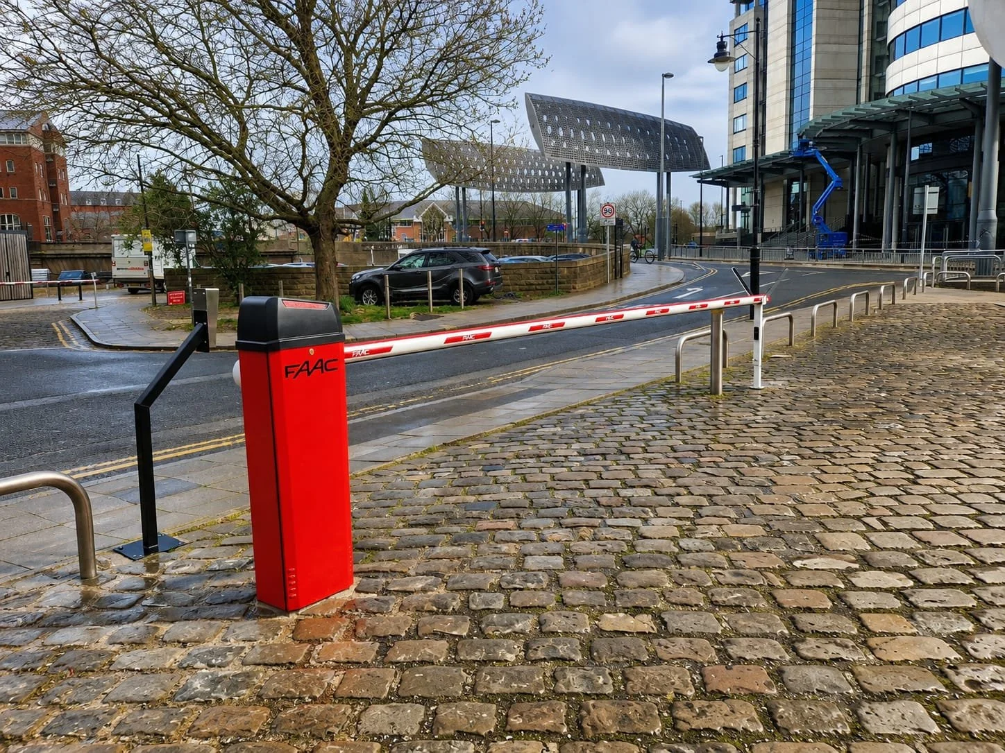 A street scene with a red barrier gate marked 'FAAC' and a raised white and red arm gate. There is cobblestone pavement, a leafless tree, parked cars, modern glass buildings, and a cloudy sky.