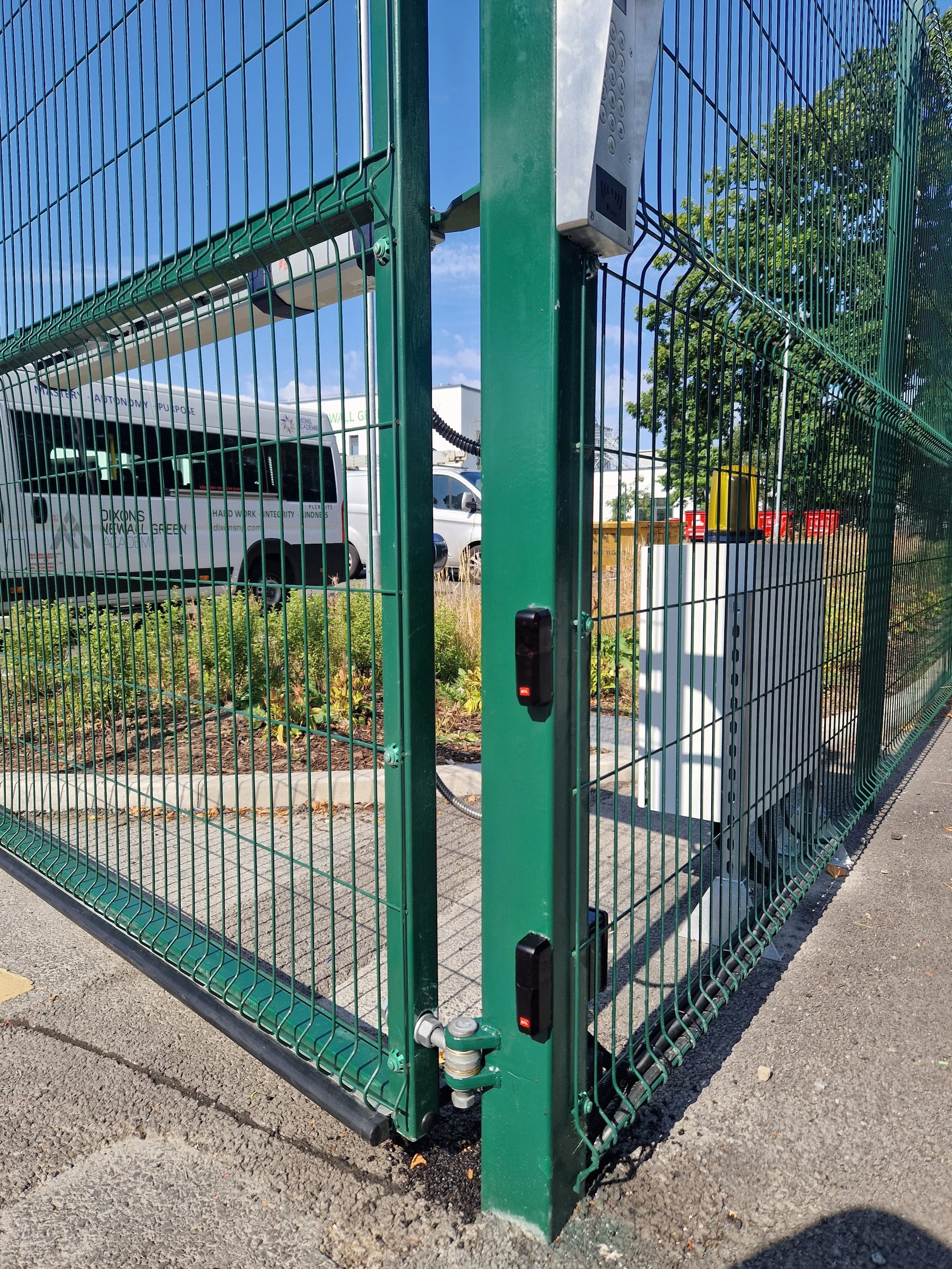 A green metal fence with electrical equipment attached, surrounding a small area with plants, a sidewalk, and a white van in the background under a clear blue sky.