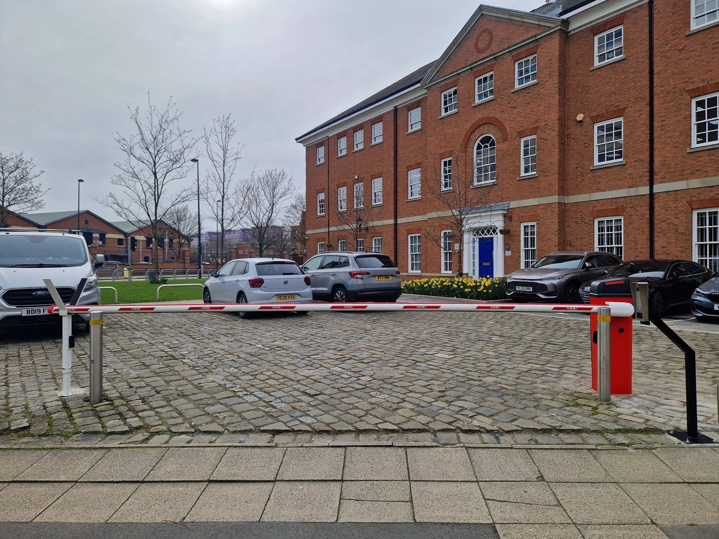 A parking lot in front of red brick buildings, with several parked cars and leafless trees. The sky is overcast.