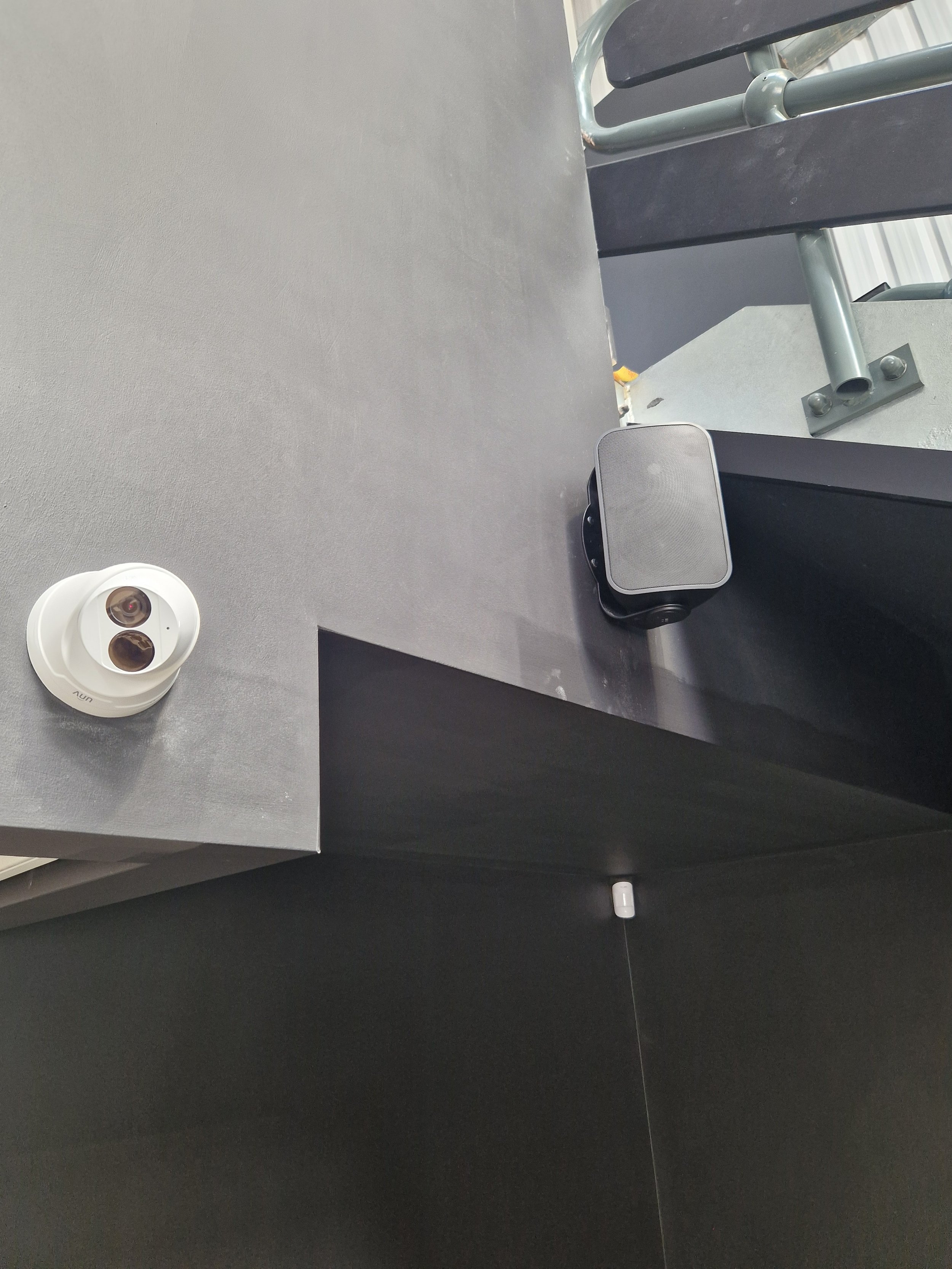 Indoor setting with a gray table, a security camera on the left, a black speaker on the table, a gray metal rod attached to the structure, and part of a black structure in the background.