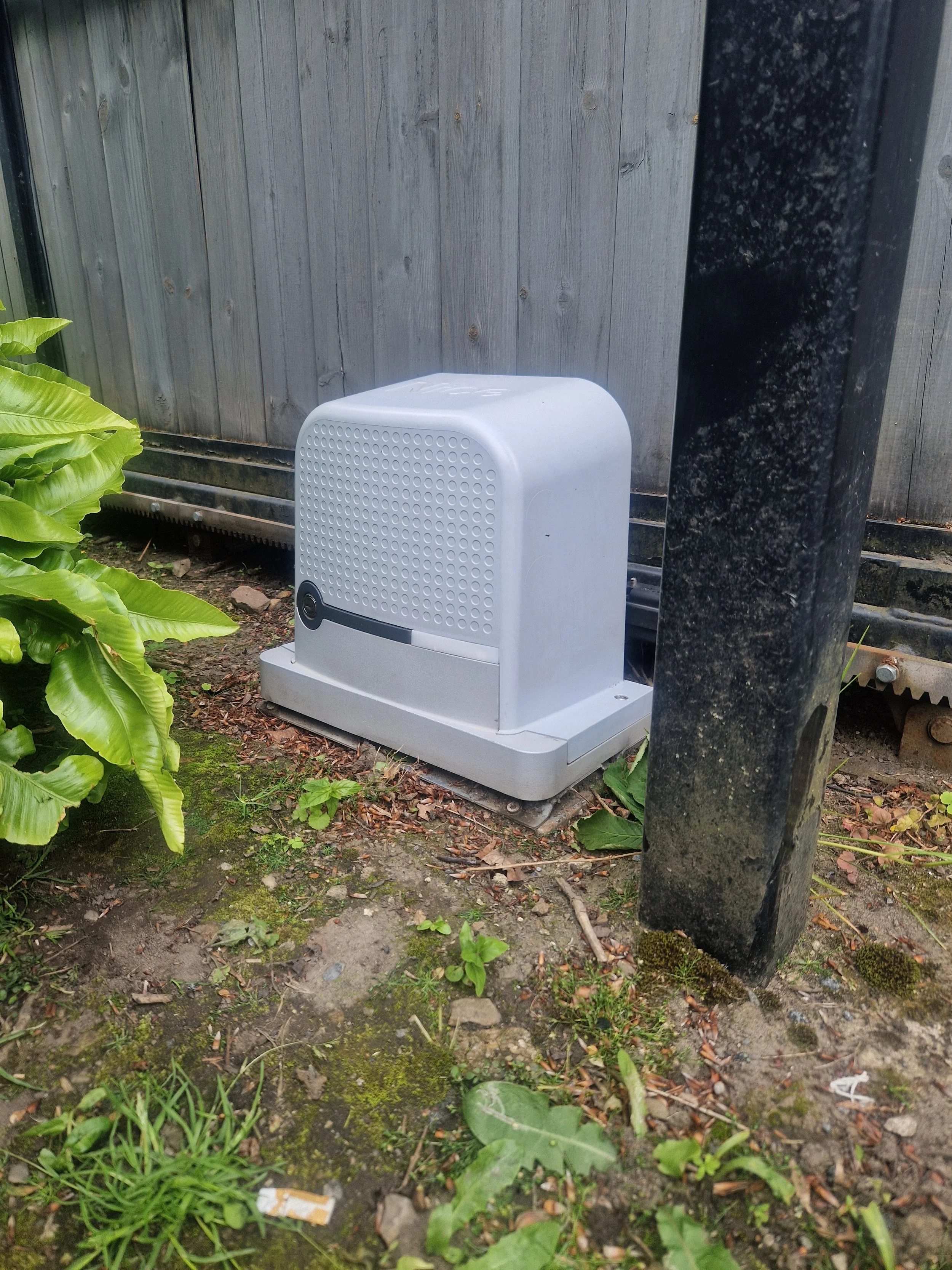An outdoor scene showing a white air purifier on the ground next to a black pole, with a wooden fence in the background and various green plants and weeds in the soil.