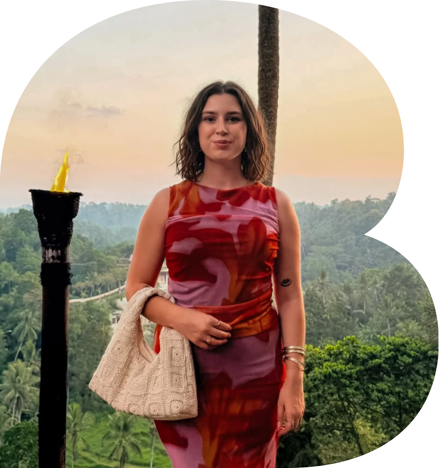 A woman with shoulder-length wavy brown hair standing outdoors during sunset. She is wearing a sleeveless, multi-colored dress with red, pink, and orange hues, and is holding a beige knitted bag. There is a tattoo of a black heart shape on her right upper arm, and she has rings on her fingers and bracelets on her left wrist. A torch with a yellow flame is seen to her left, and a tall palm tree is behind her. The background includes lush green trees and a distant view of a hilly landscape.