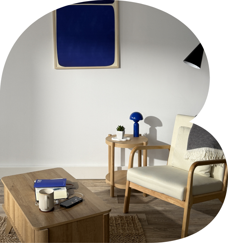 Cozy living room corner with a wooden armchair, a round side table with a lamp and plant, a coffee table with books, a mug, and electronic device, a framed blue abstract art piece on the white wall, and a black wall-mounted lamp.