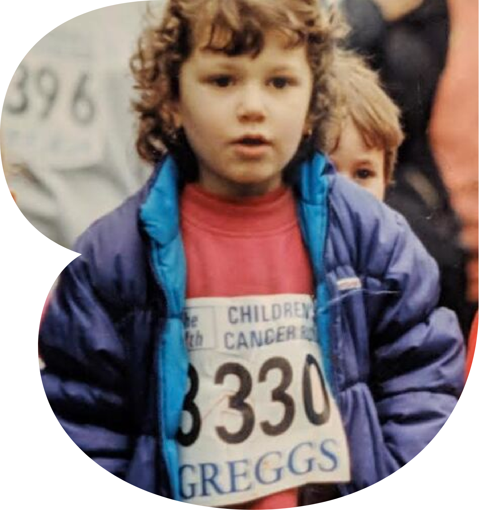 A young boy with brown curly hair wearing a red shirt and a purple jacket, standing among others and holding a race bib with the number 2330 on it.