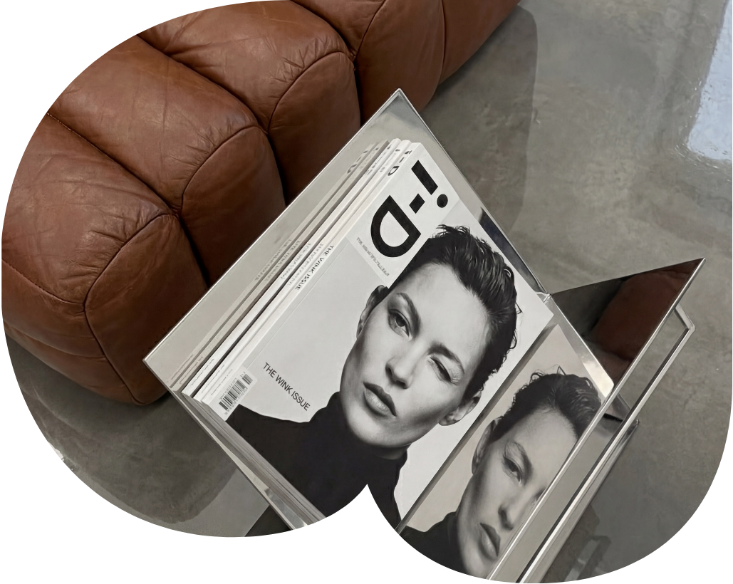 A magazine rack holding copies of a magazine with a black and white cover photo of a woman making a winking expression. The rack is next to a brown leather couch on a concrete floor.