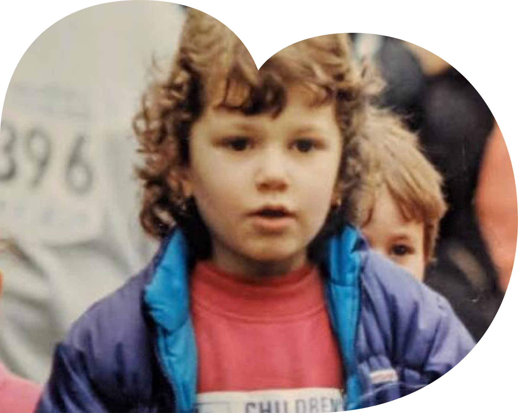 Young boy with curly hair wearing a blue jacket and red shirt, standing in front of a white background with a blurred logo or text.