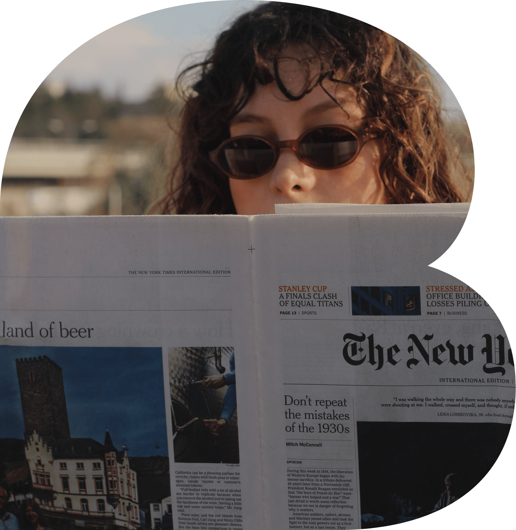 Person with curly hair and sunglasses reading a newspaper outdoors.