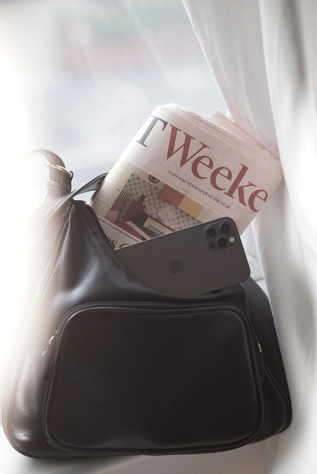 A black leather bag with a newspaper, a smartphone, and a white curtain nearby.